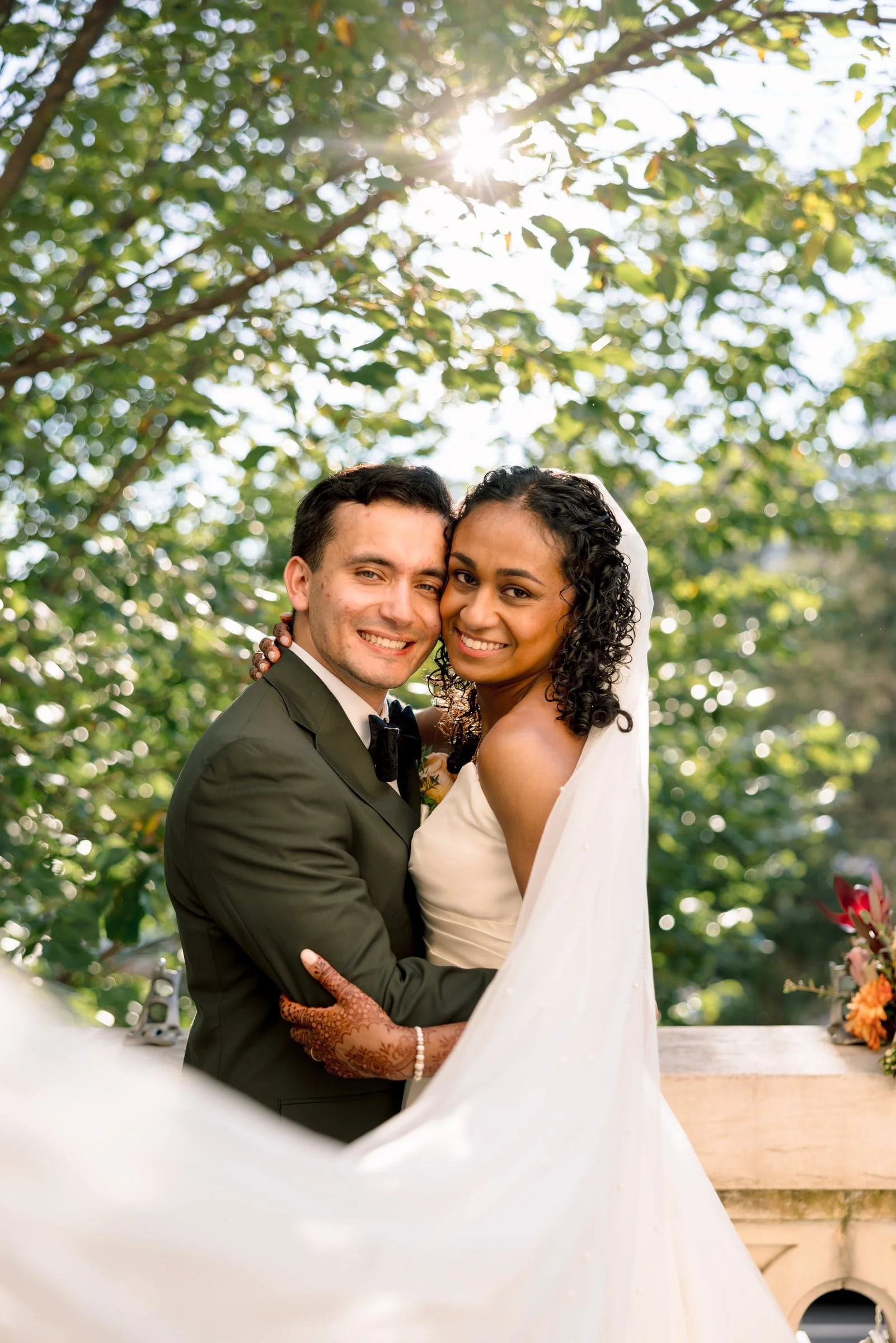 A smiling couple, dressed in wedding attire, embracing outdoors with green trees and sunlight in the background.