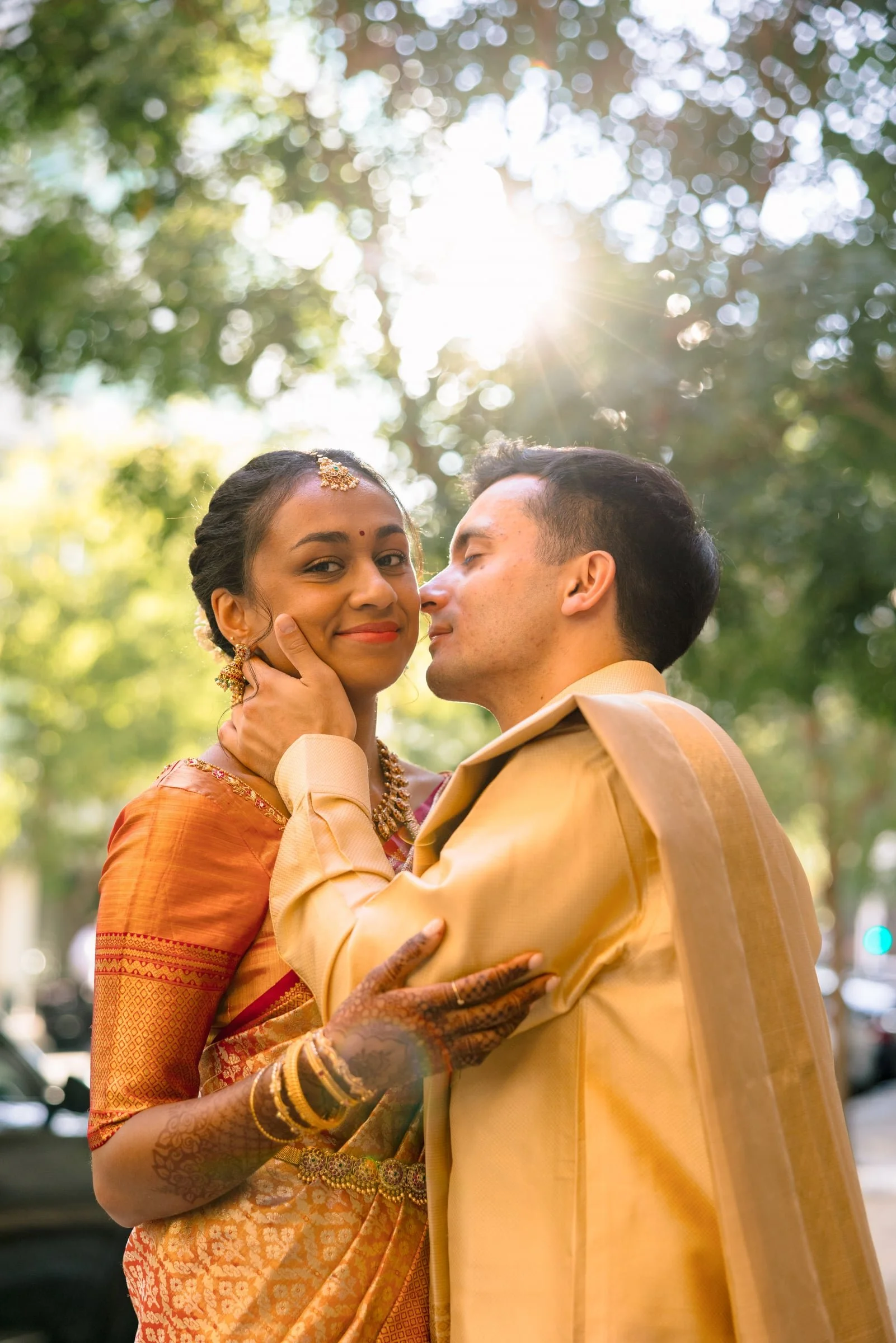 An Indian couple dressed in traditional attire, outdoors on a sunny day surrounded by trees and greenery, sharing an intimate moment.