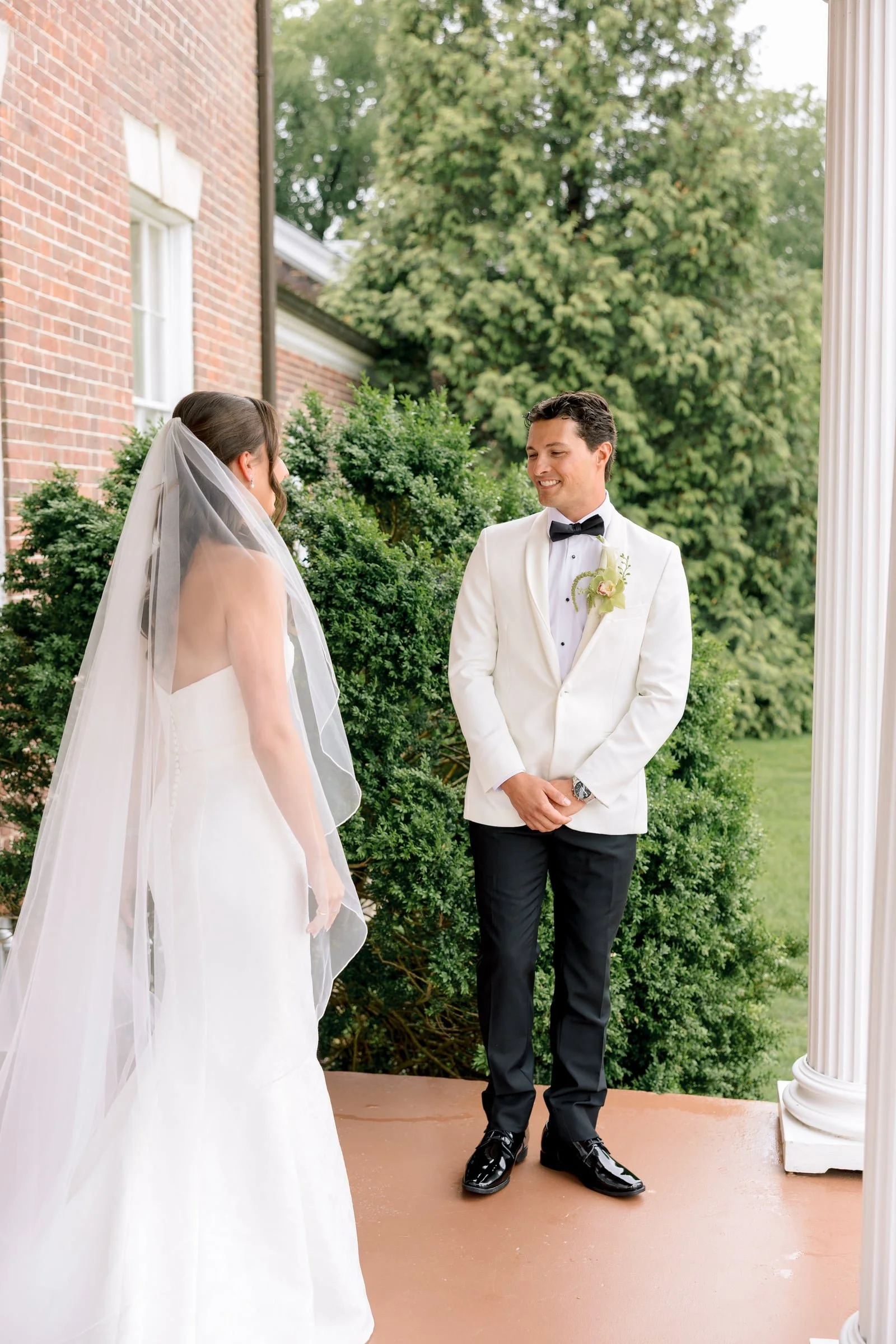 A bride and groom stand outside on a porch, facing each other. The bride wears a white wedding gown with a veil, and the groom wears a white tuxedo jacket with black pants, a black bow tie, and a boutonniere. They are smiling at each other with green