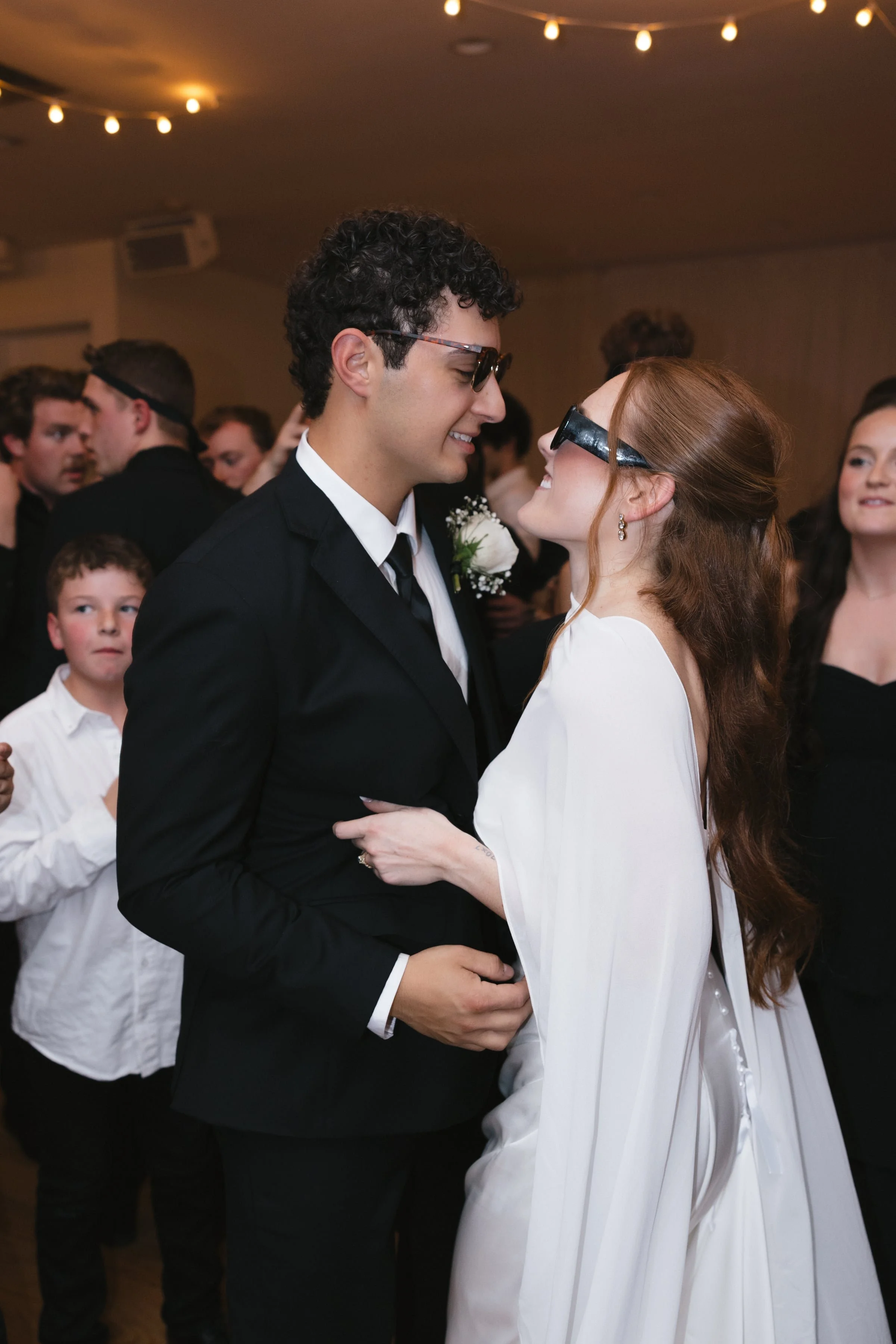 A couple dancing closely at a formal event, smiling at each other, with guests in the background. The man has curly hair, glasses, and is wearing a black suit with a white shirt and a boutonniere. The woman has long red hair, earrings, and is wearing