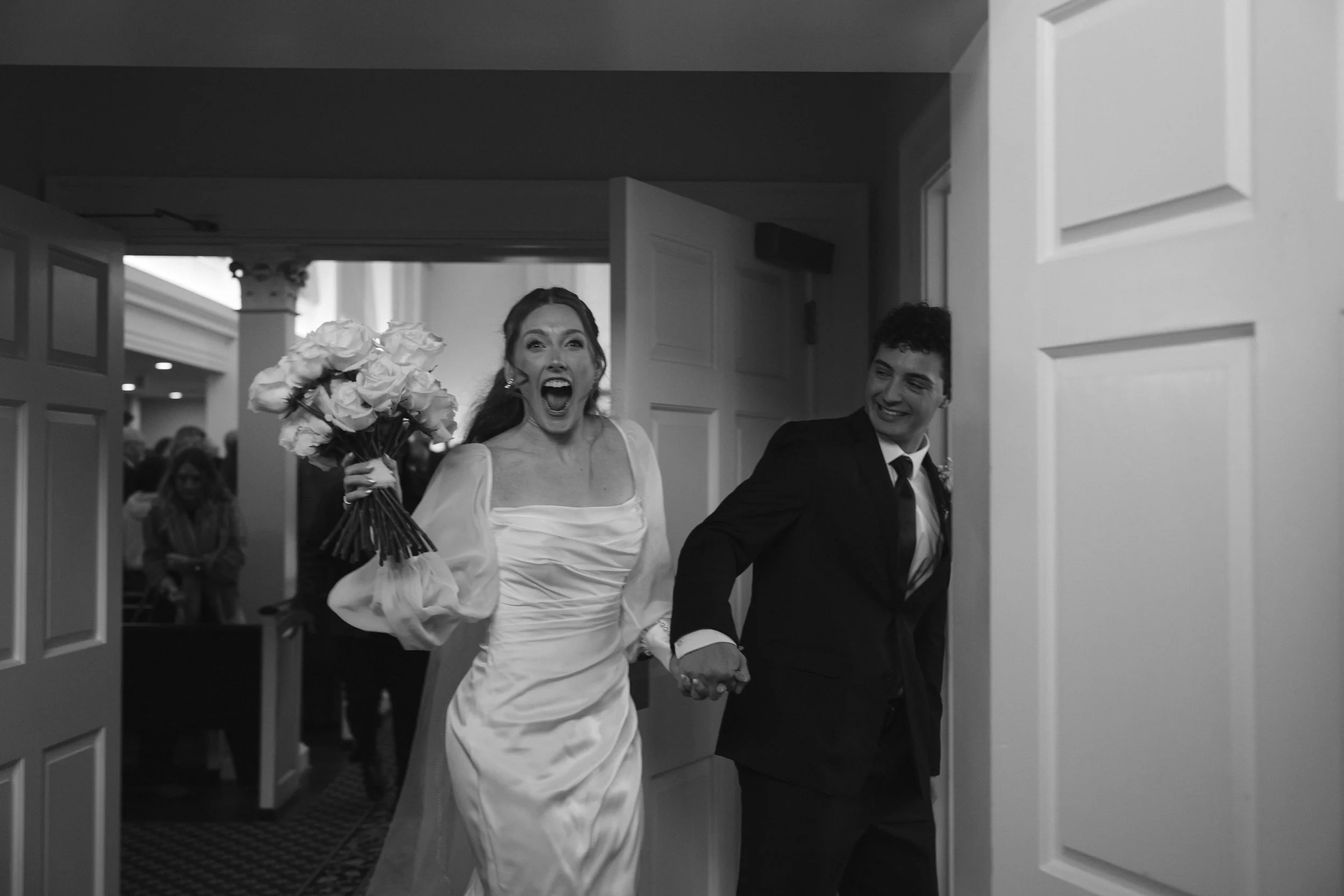 A woman in a white wedding dress holding a bouquet of roses, smiling and laughing, as she walks arm-in-arm with a man in a dark suit, both entering a room through a door, with onlookers in the background.