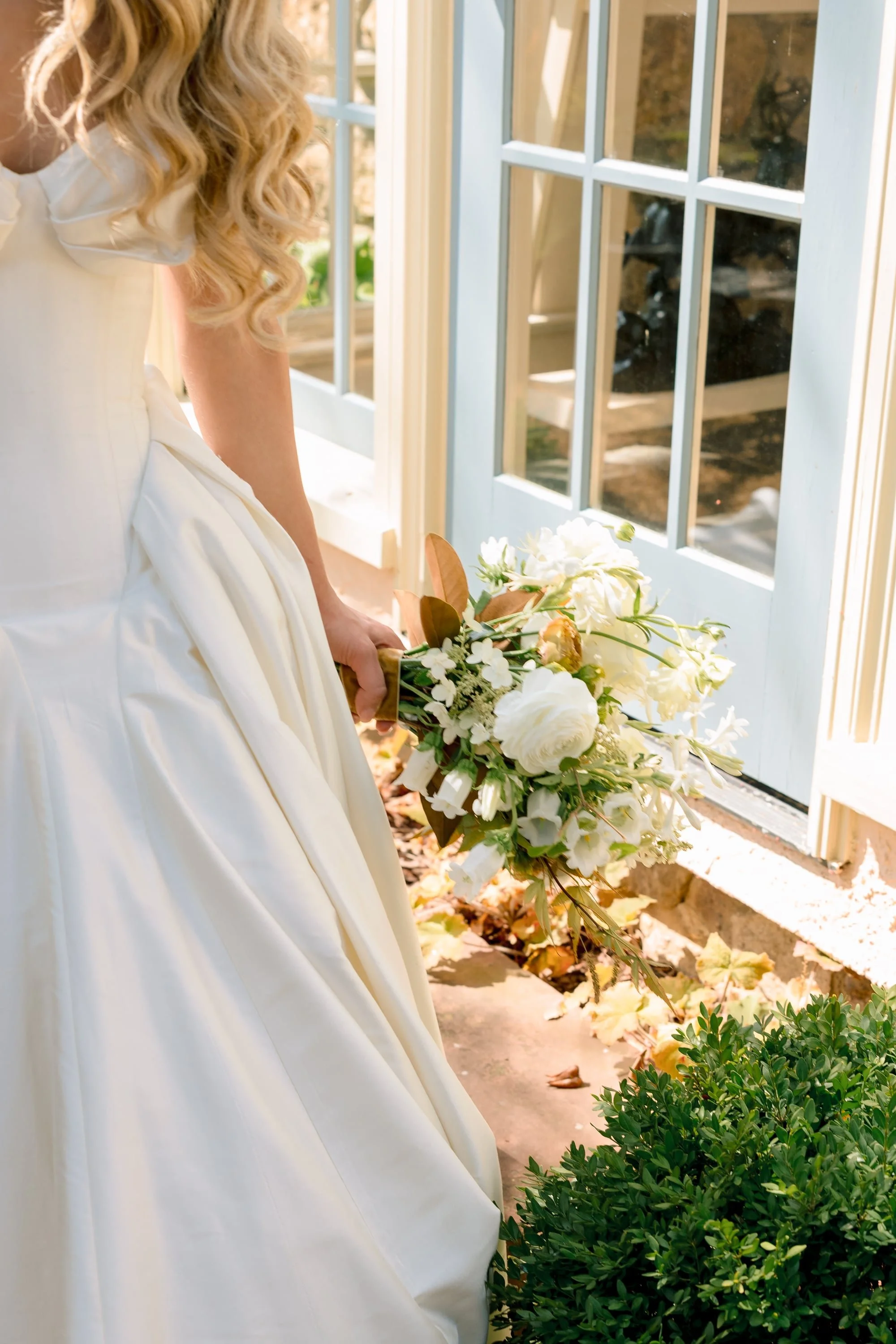 A woman in a white dress holding a bouquet of white flowers standing near a glass door with a wooden frame, sunlight streaming in, and autumn leaves on the ground.