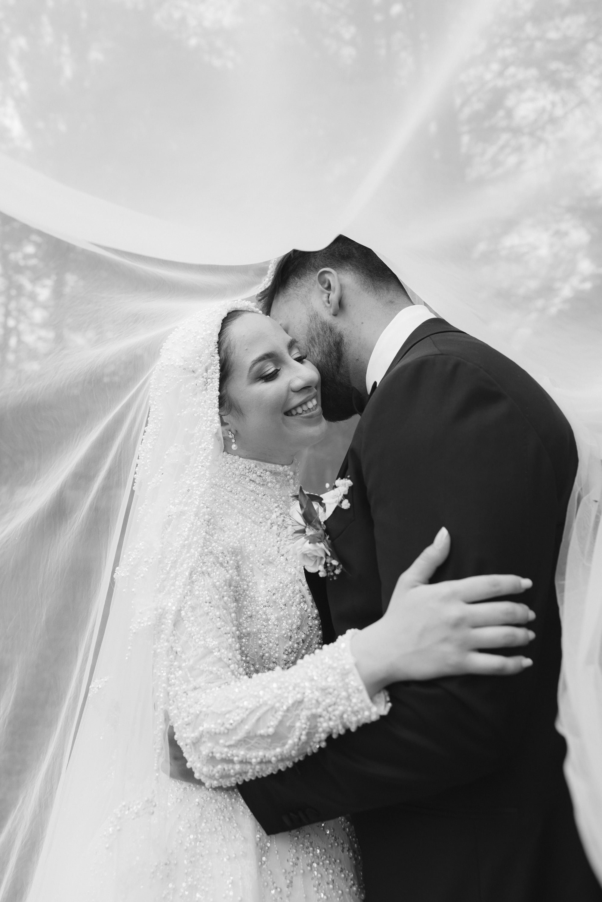 A black and white photo of a bride and groom kissing, with the bride wearing a beaded wedding dress and veil, and the groom in a suit and tie, embracing each other closely.