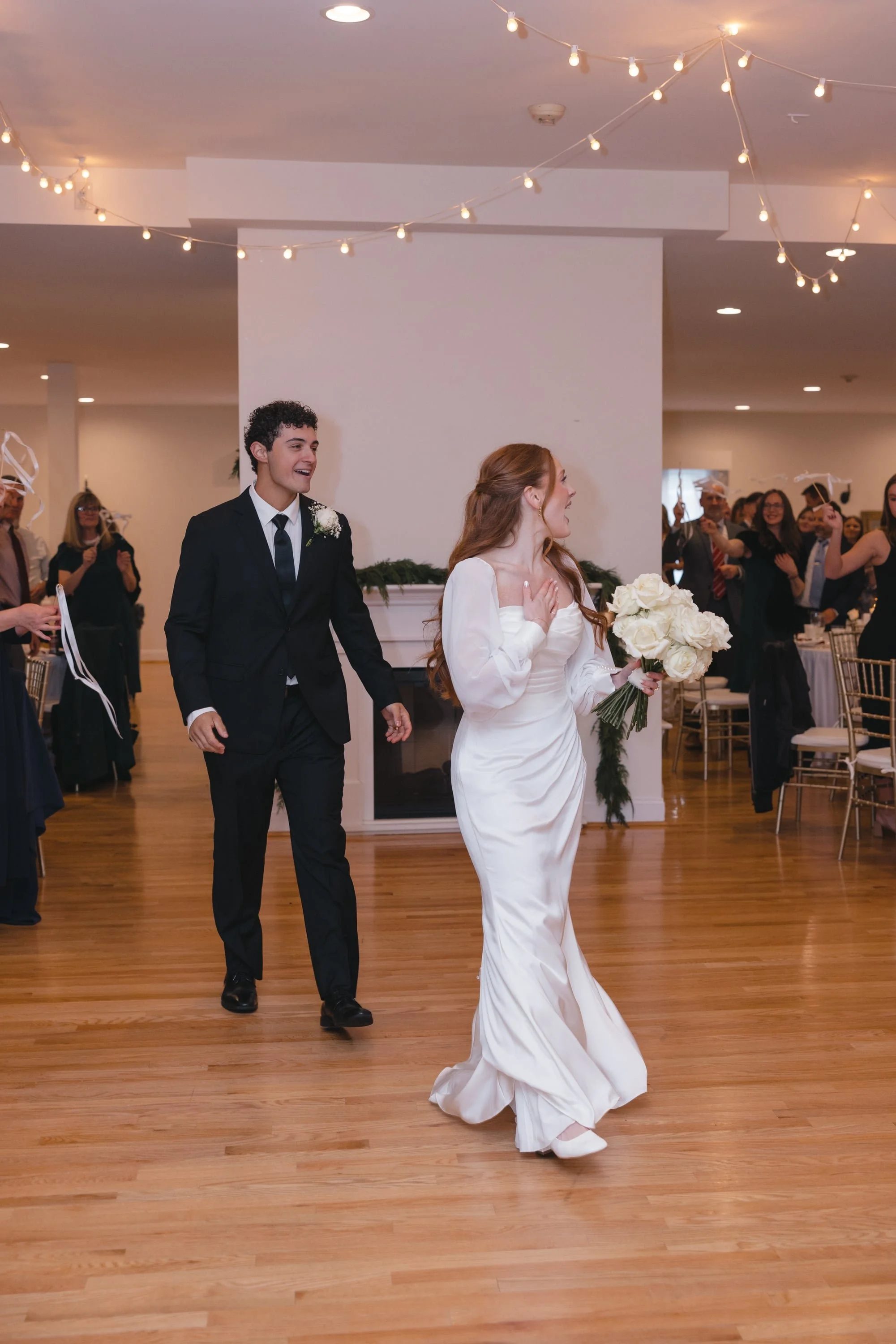 A bride and groom holding hands and smiling during their wedding reception, with guests clapping and celebrating in the background.