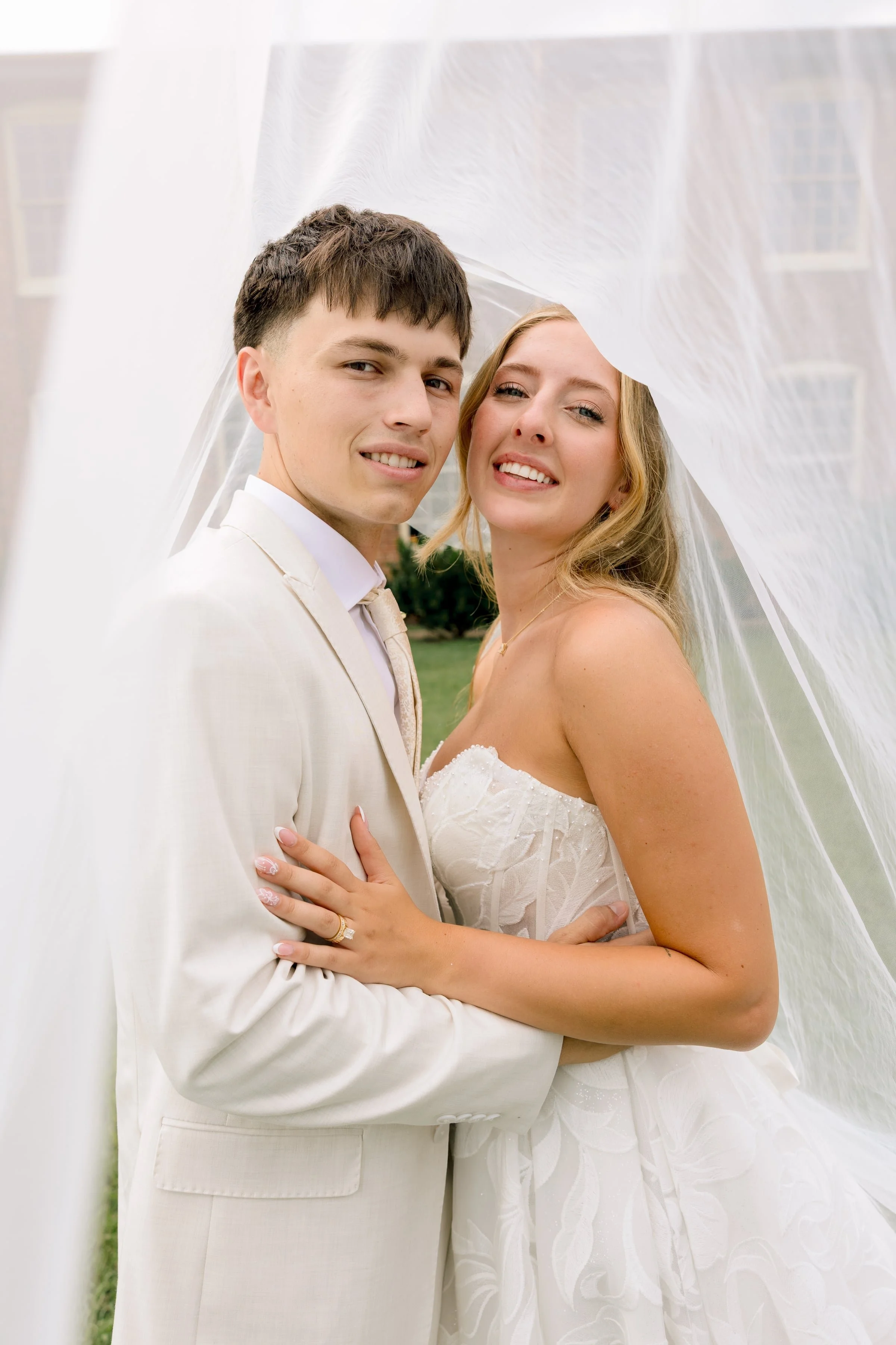 A newlywed couple standing close together, both smiling, under a white veil during their wedding day, outdoors in front of a building with large windows.