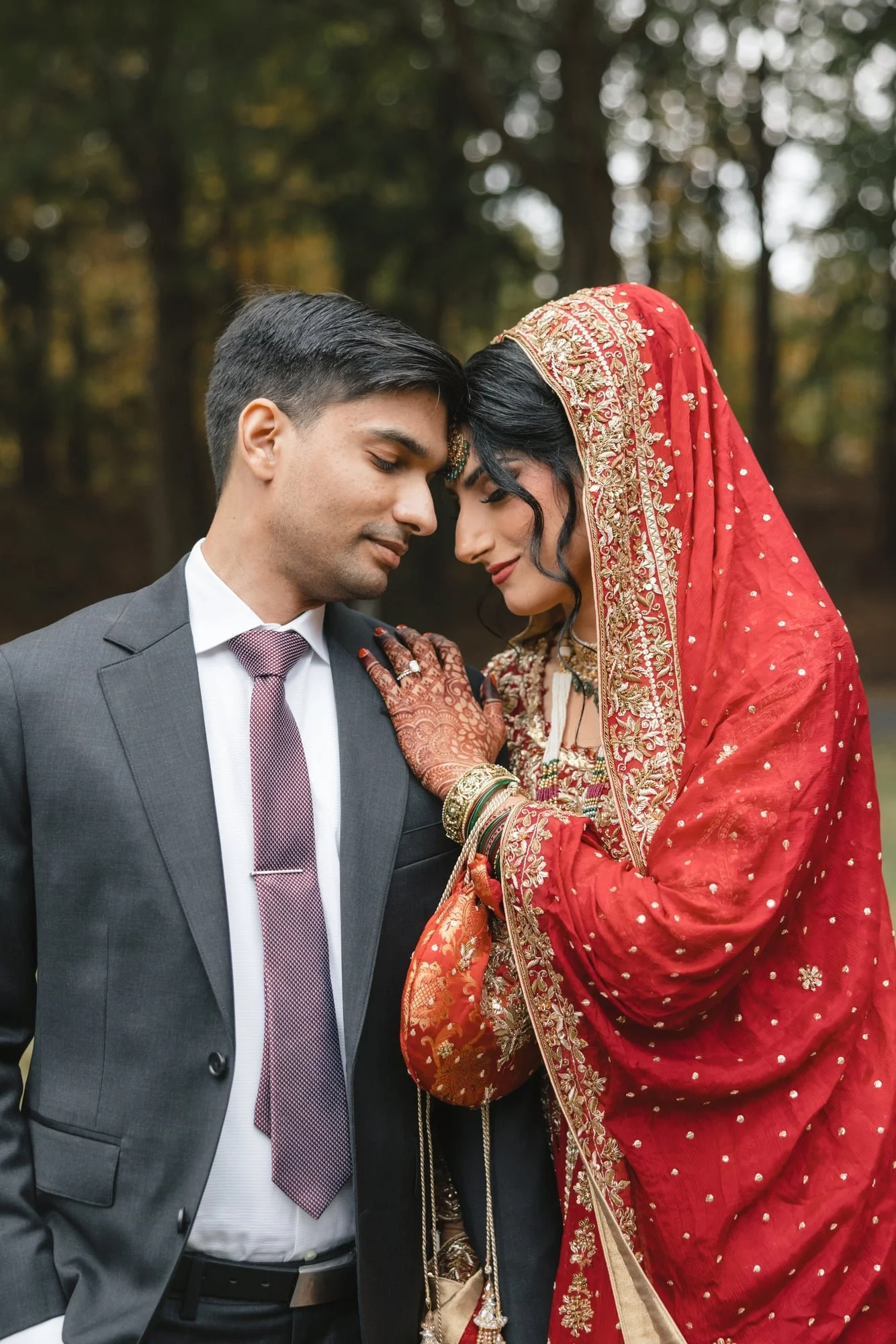 A couple posed together outdoors, with their foreheads touching, the woman wearing traditional Indian attire and jewelry, and the man dressed in a suit, with blurred trees in the background.