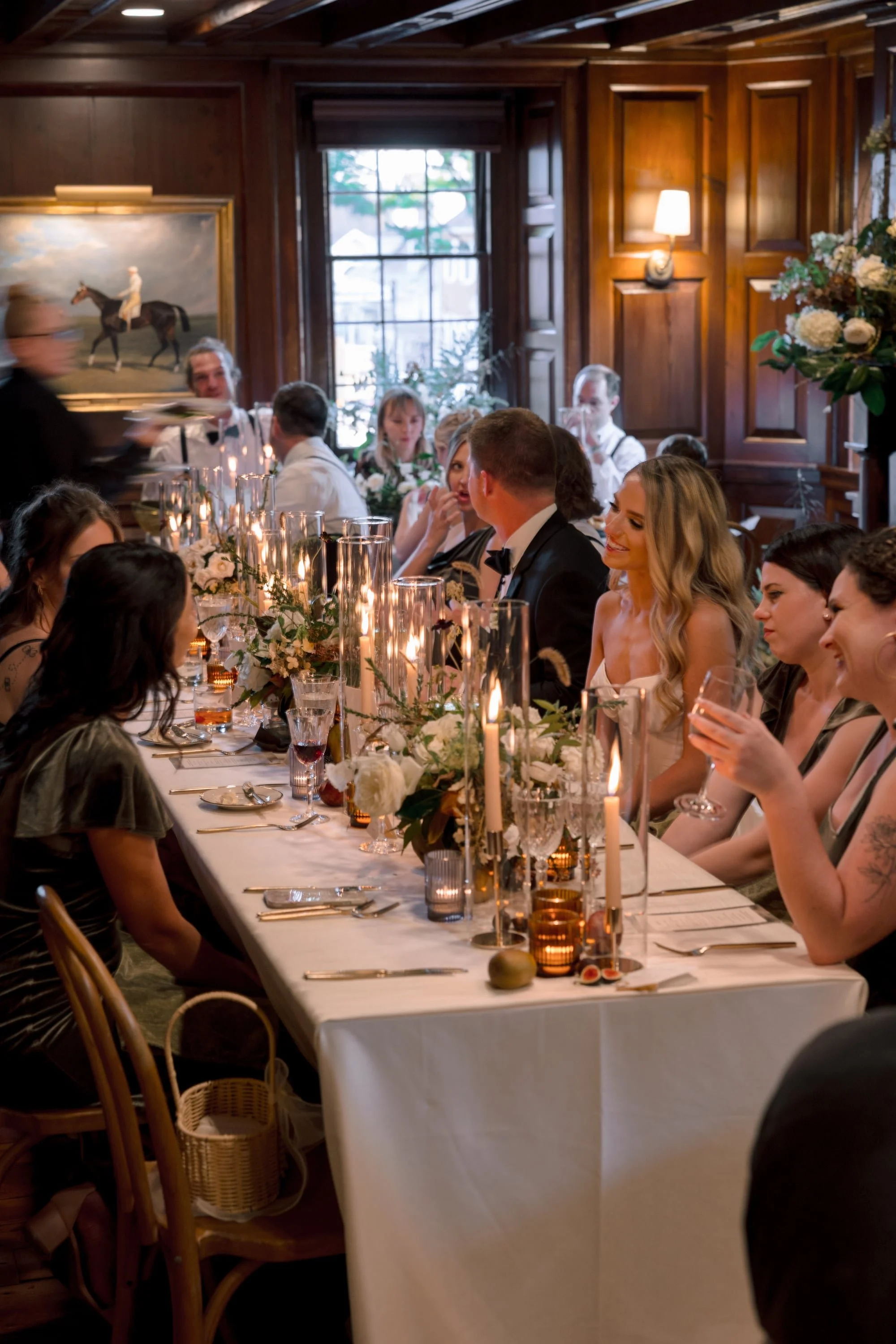 People sitting at a long dinner table with white tablecloth, candles, and floral centerpieces in a wood-paneled room, attending a formal event or celebration.