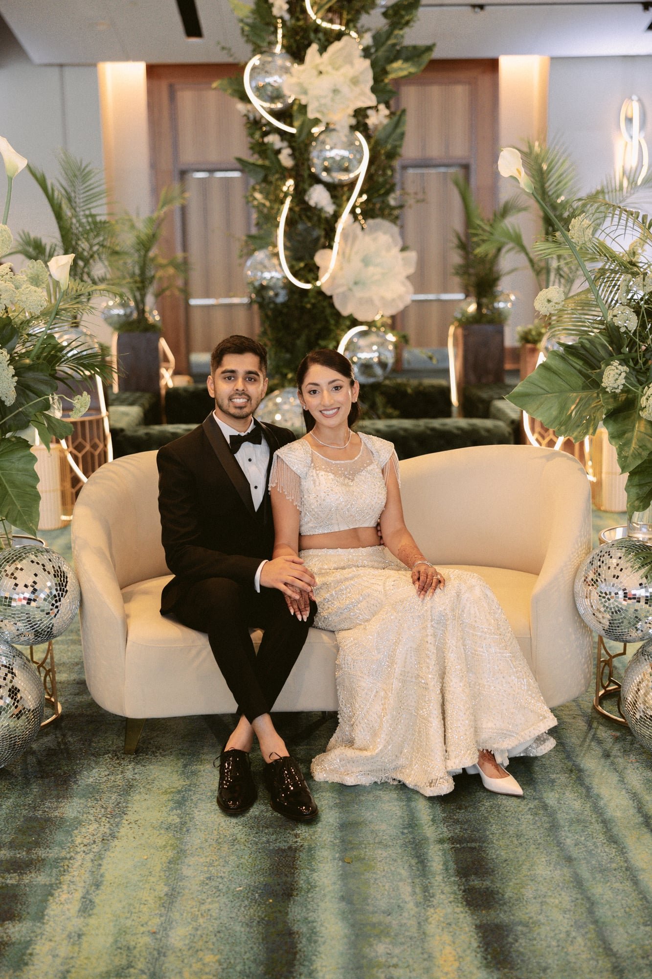 A couple dressed in formal attire sitting on a cream-colored sofa, celebrating their wedding. The man is wearing a black tuxedo with a bowtie, and the woman is in a white wedding dress with intricate beadwork. They are in a decorated indoor venue wit