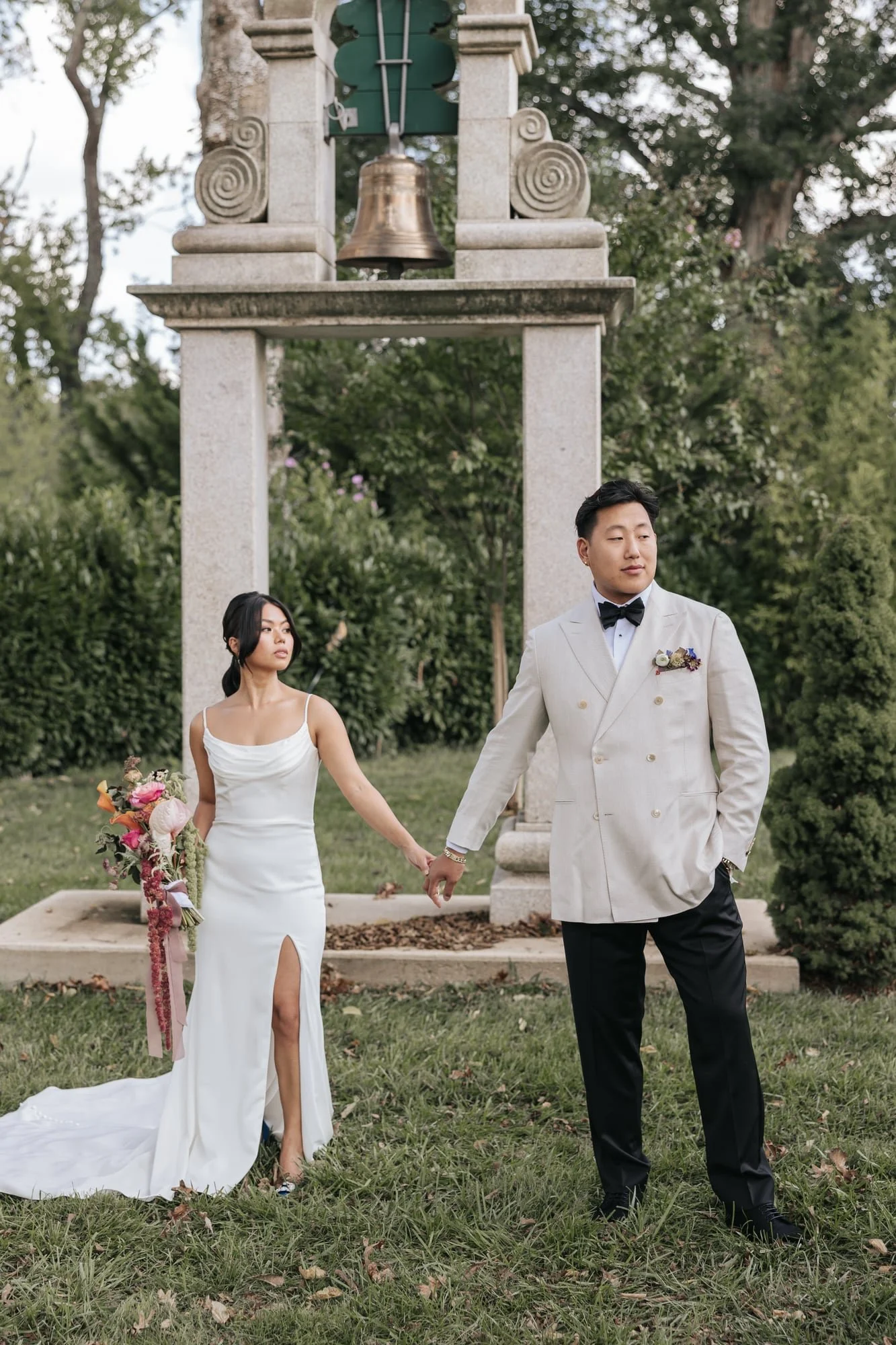 A bride and groom holding hands outdoors, standing in front of a stone arch with a bell. The bride wears a white dress with a slit, and the groom wears a light-colored tuxedo with a black bow tie. They are surrounded by trees and greenery.