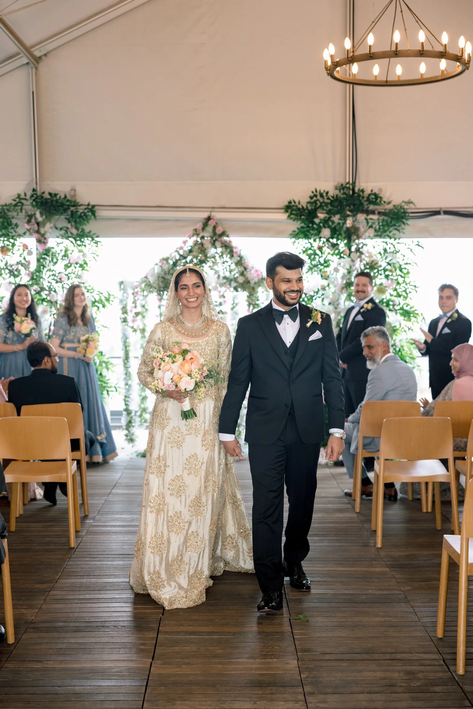 Happy bride and groom walking down the aisle at a wedding ceremony. The bride is wearing a cream-colored dress with gold embroidery and a veil, holding a bouquet of pink and cream flowers. The groom is dressed in a black tuxedo with a bow tie. Guests