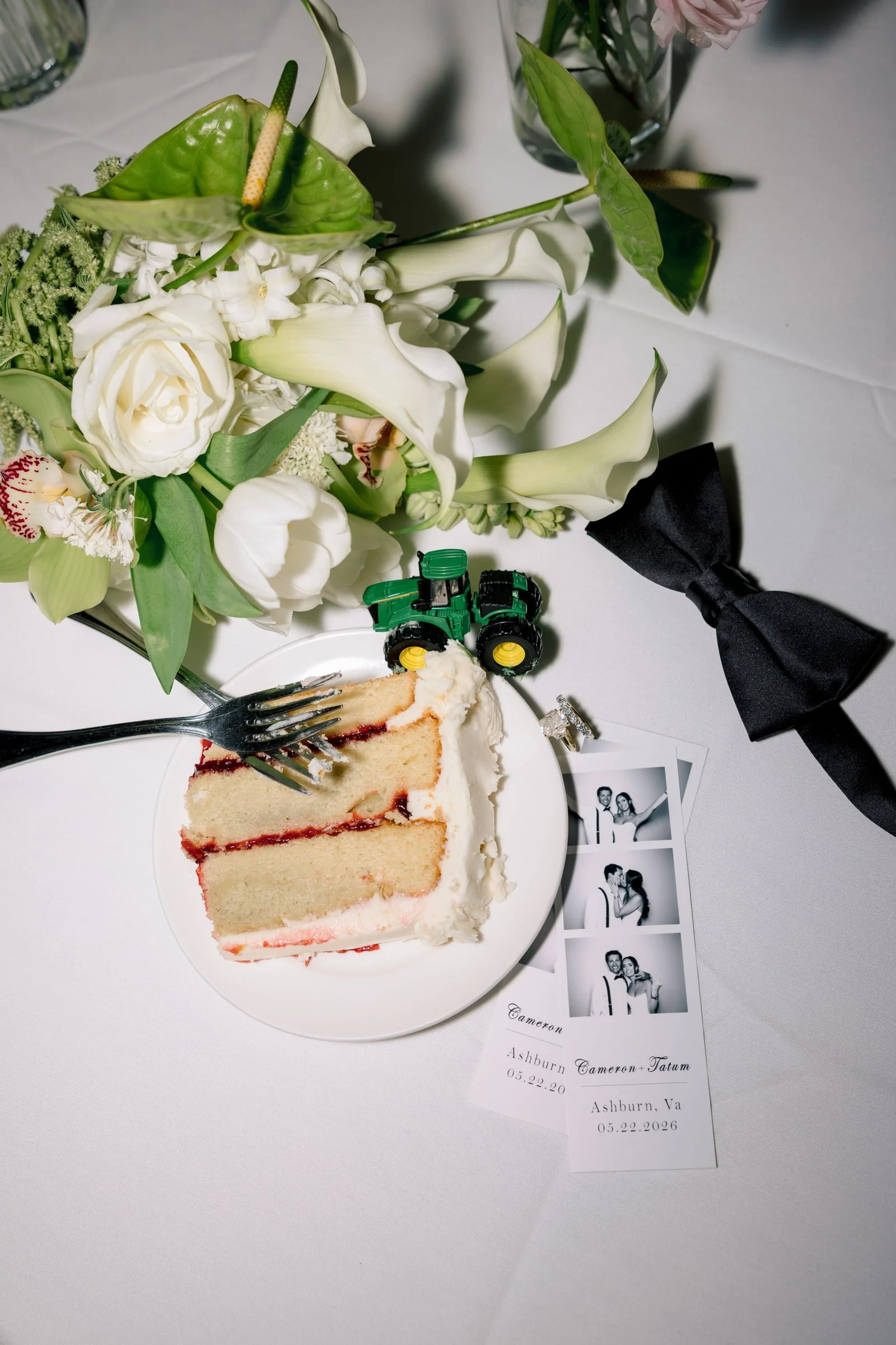 A slice of wedding cake with white frosting and red filling on a white plate, a fork, a bouquet of white lilies and other flowers, a small toy tractor, and photo booth pictures on a white table with a black ribbon and wedding cards.