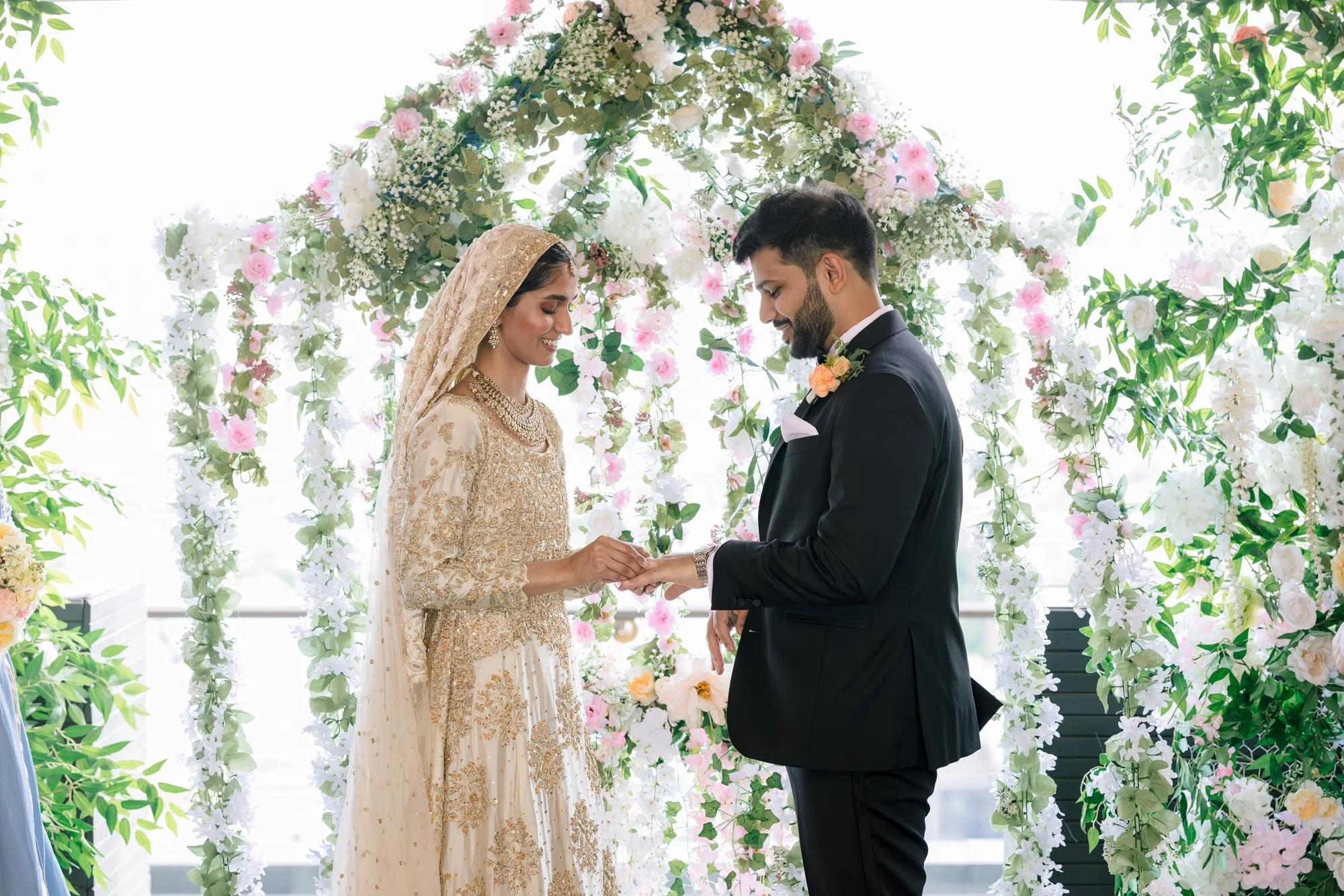 Couple getting married standing under a floral wedding arch, with the bride wearing a gold embroidered traditional dress and a dupatta, and the groom in a black suit with a boutonniere, holding hands and smiling.