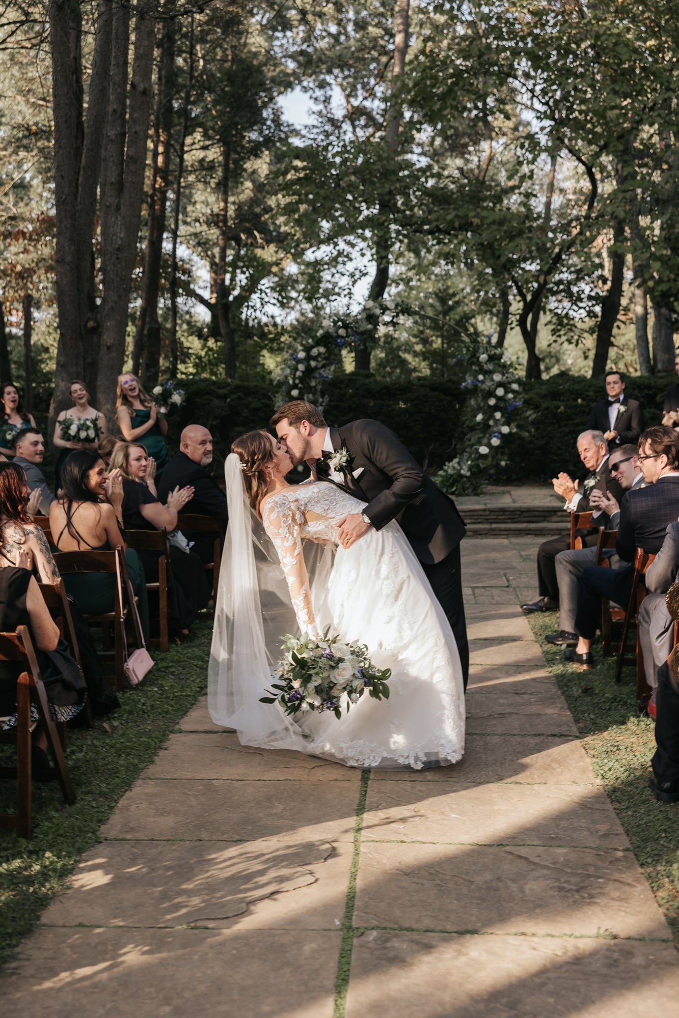 A bride and groom sharing a kiss during their outdoor wedding ceremony, with guests seated on either side of the aisle, surrounded by trees and floral decorations.