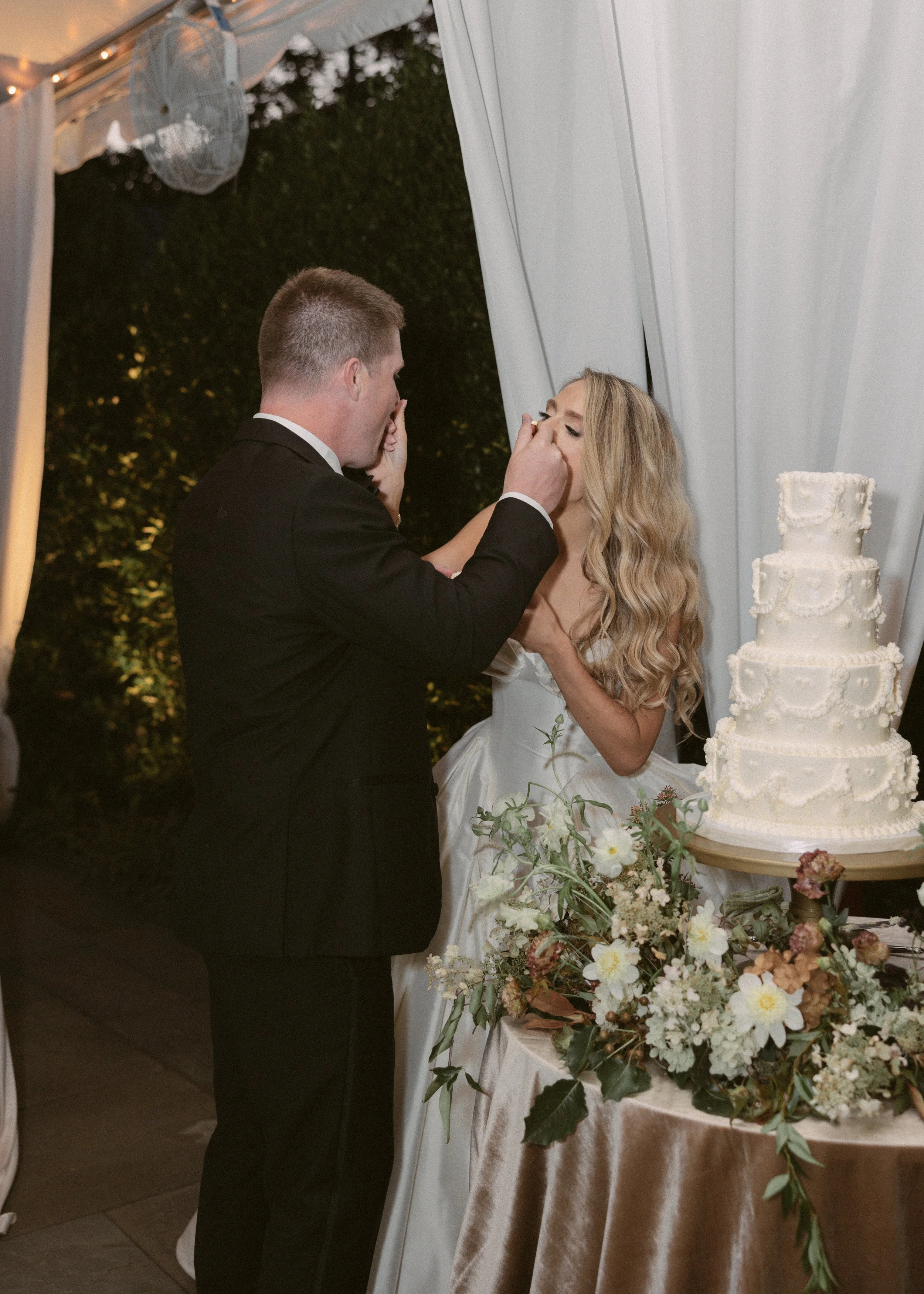 A bride and groom at their wedding cut the wedding cake together under a tent decorated with white drapes and fairy lights. The bride has long, wavy blonde hair and is wearing a white wedding dress. The groom has short, dark hair and is wearing a bla