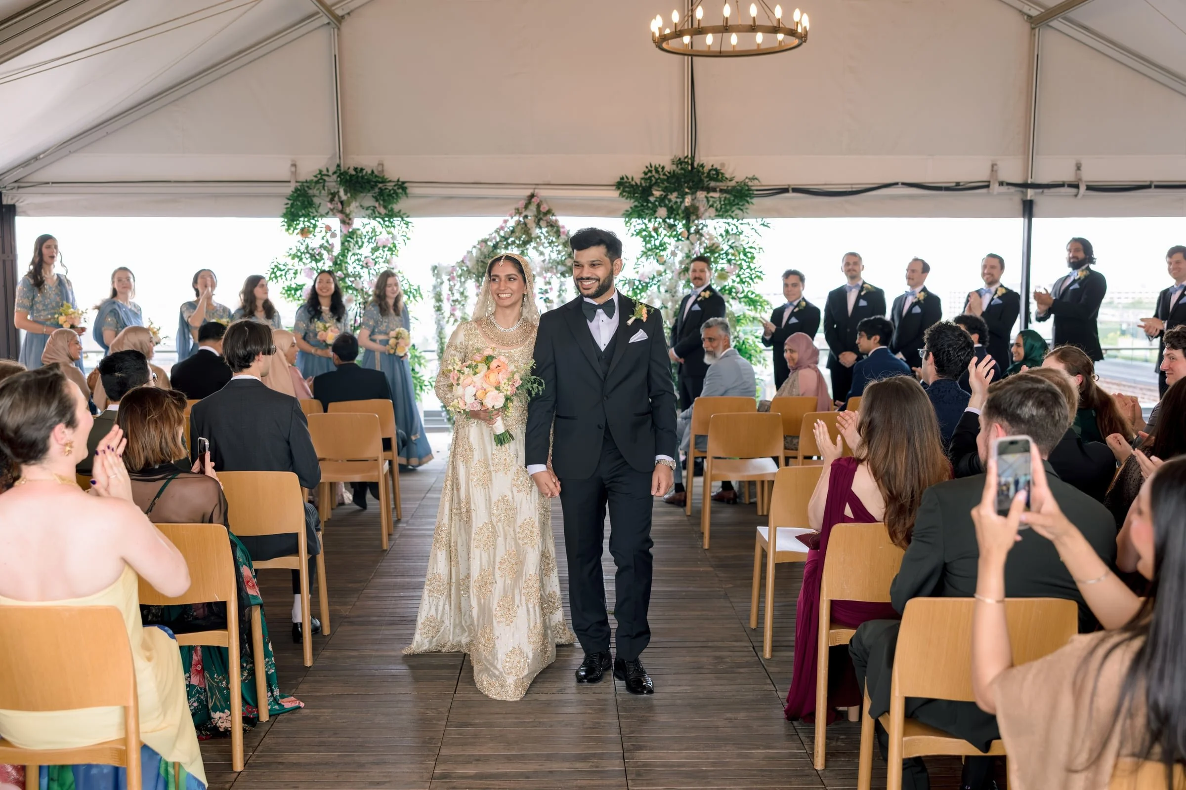 A bride and groom walking down the aisle at a wedding ceremony, surrounded by seated guests and wedding party members with a floral background.
