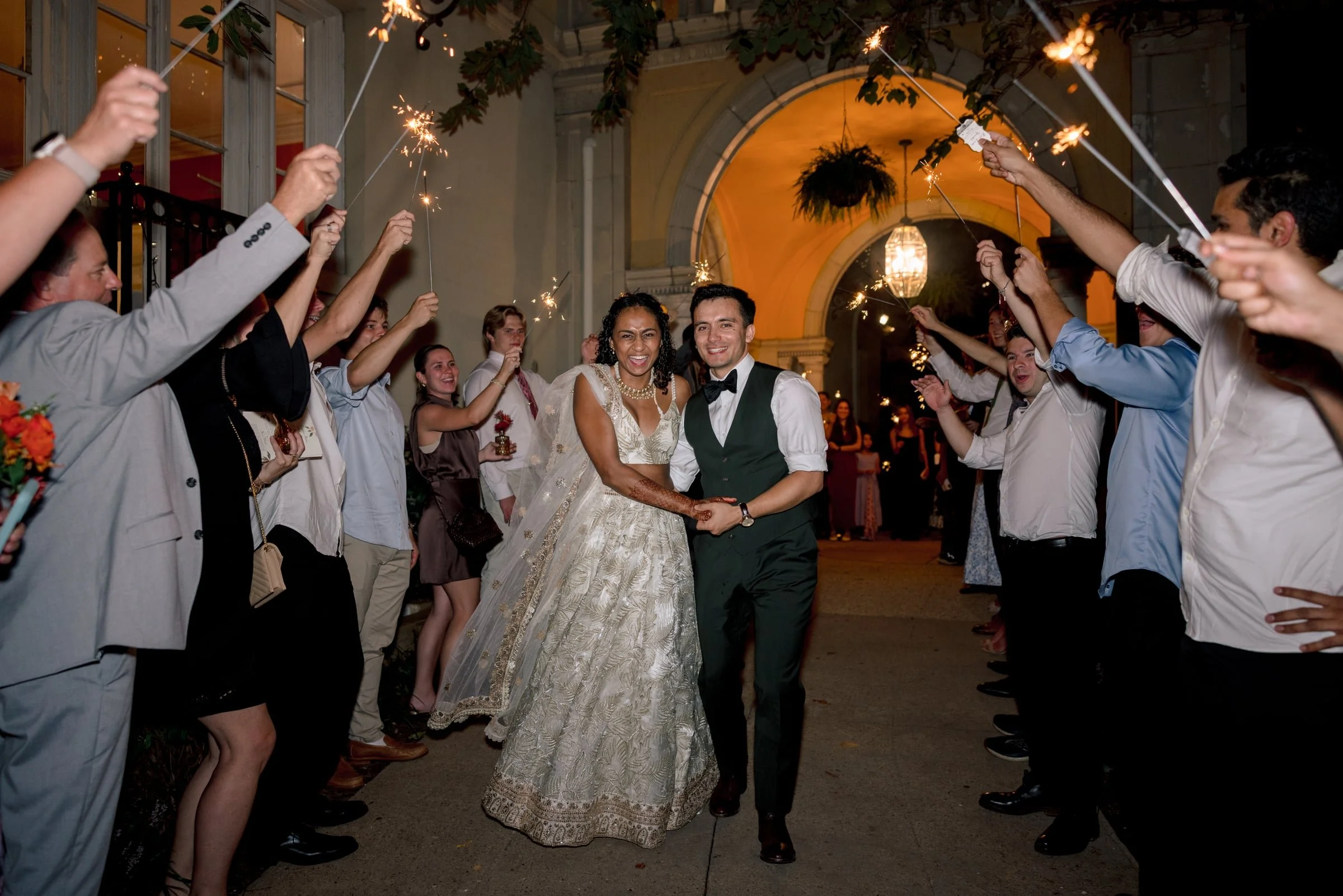 A newlywed couple walking through a line of friends and family holding sparklers during a wedding celebration at night outside a building with arches and hanging plants.
