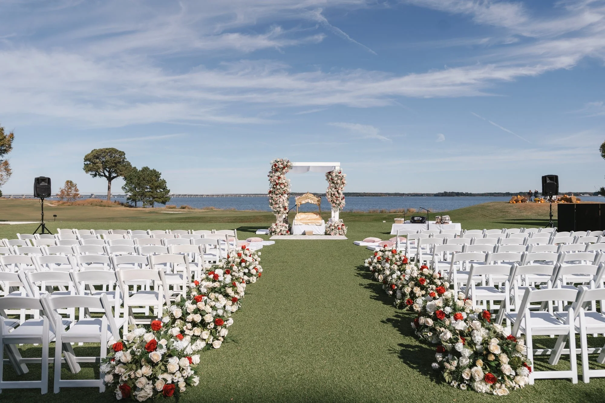 Outdoor wedding setup near a body of water with rows of white chairs, floral arrangements along the aisle, and a decorated arch for the ceremony.