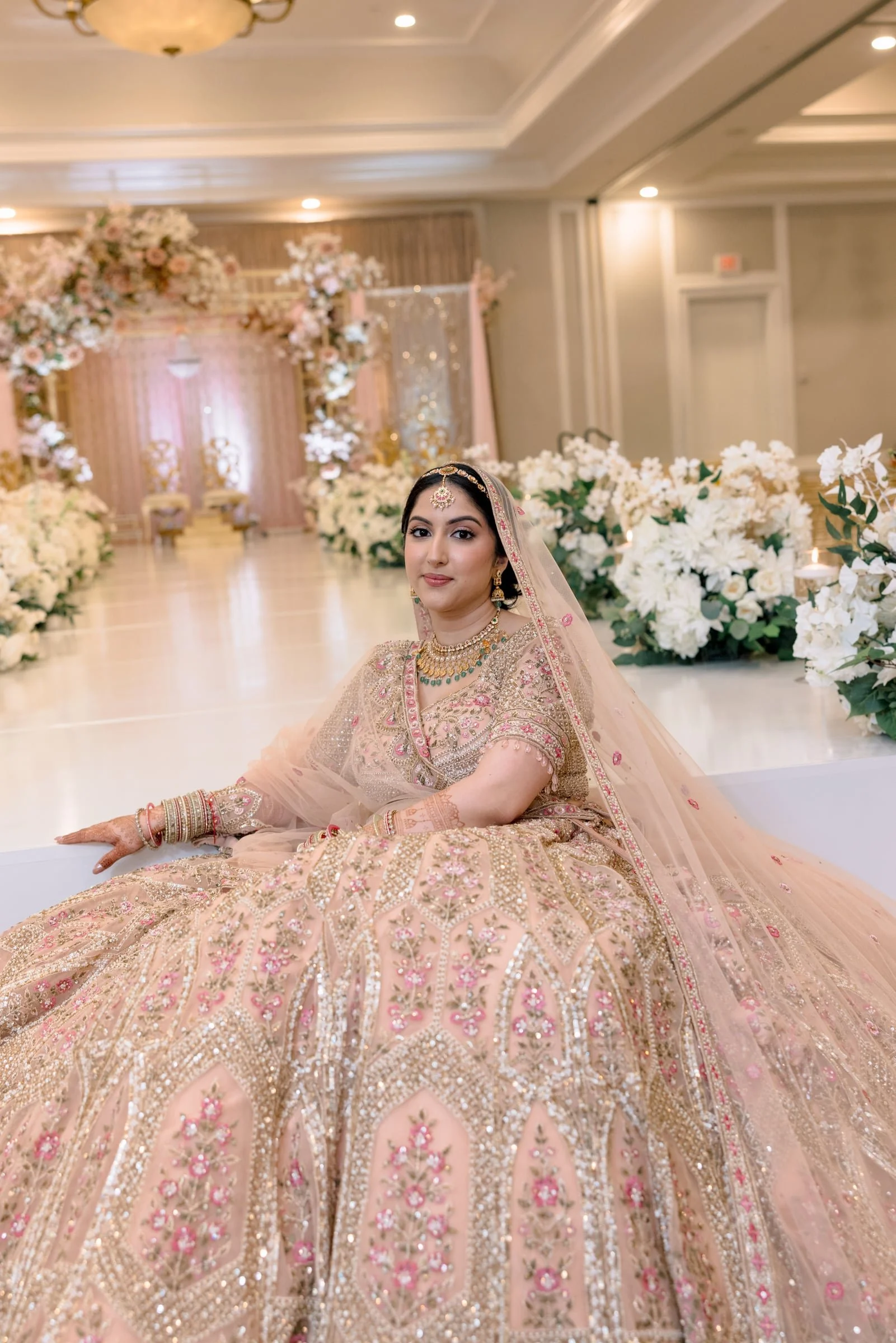A woman in traditional Indian wedding attire sitting in a decorated hall with floral arrangements and soft lighting.