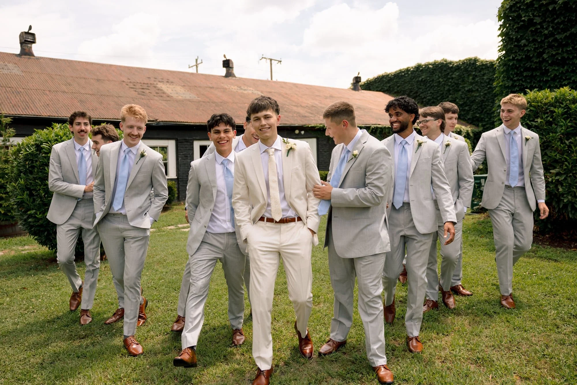 A group of young men in suits walking outdoors, smiling and laughing, during a wedding celebration.
