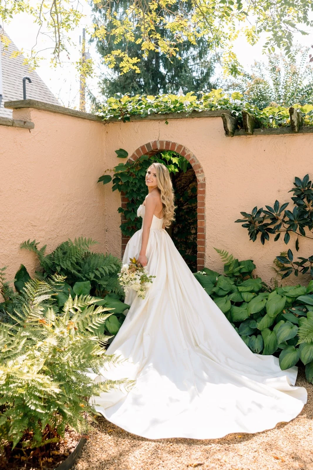Brunette bride in a strapless white gown with a long train, holding a bouquet, standing in a lush garden with green plants and a peach stucco wall with an arched brick doorway.