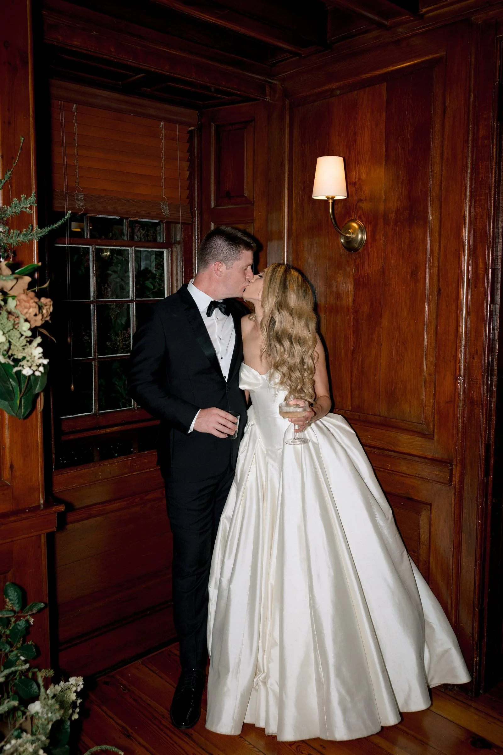A bride and groom sharing a kiss in an indoor setting with wood-paneled walls, a window with blinds, and a wall-mounted lamp.