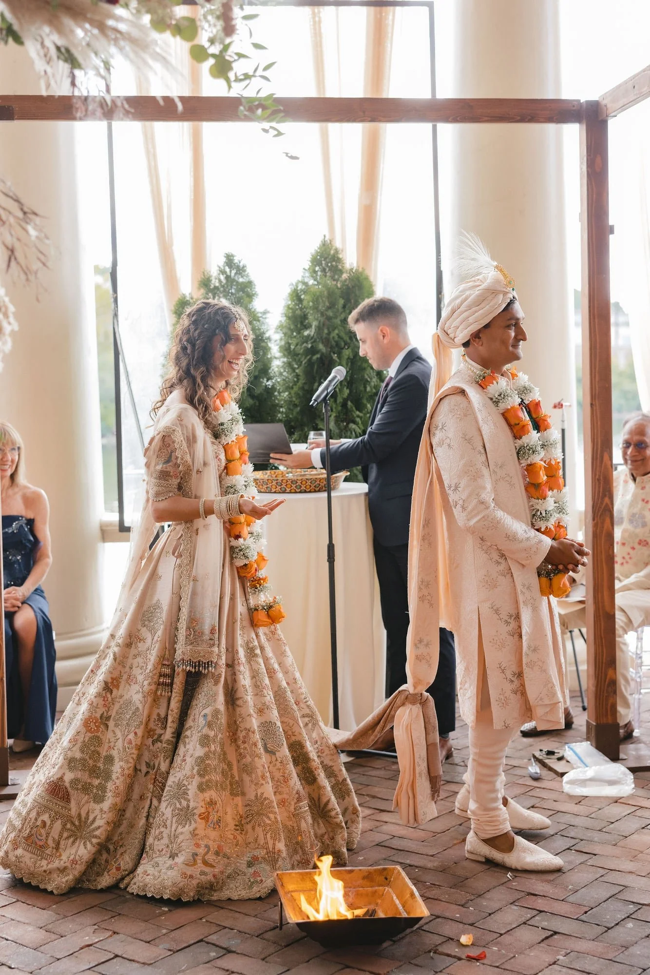 A traditional Indian wedding ceremony with a bride and groom dressed in ornate attire, with a fire ritual underway, attended by guests.