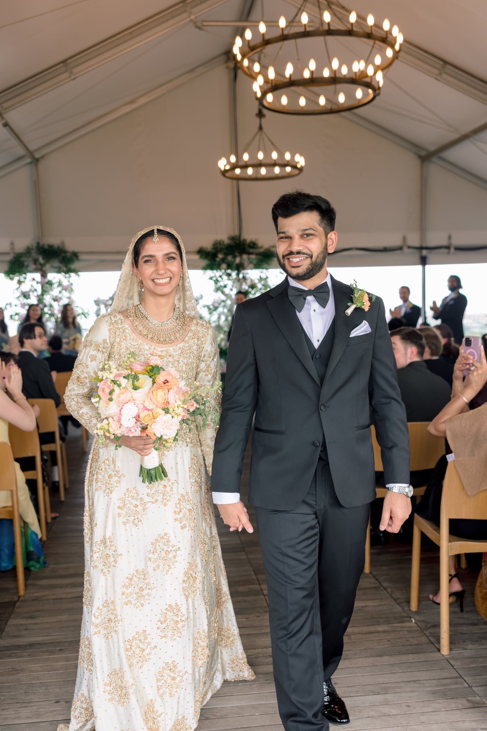 A bride and groom smiling during their wedding ceremony inside a decorated venue with chandeliers hanging from the ceiling. The bride is holding a bouquet of pink and white flowers, wearing a white and gold embroidered dress with a veil, while the gr