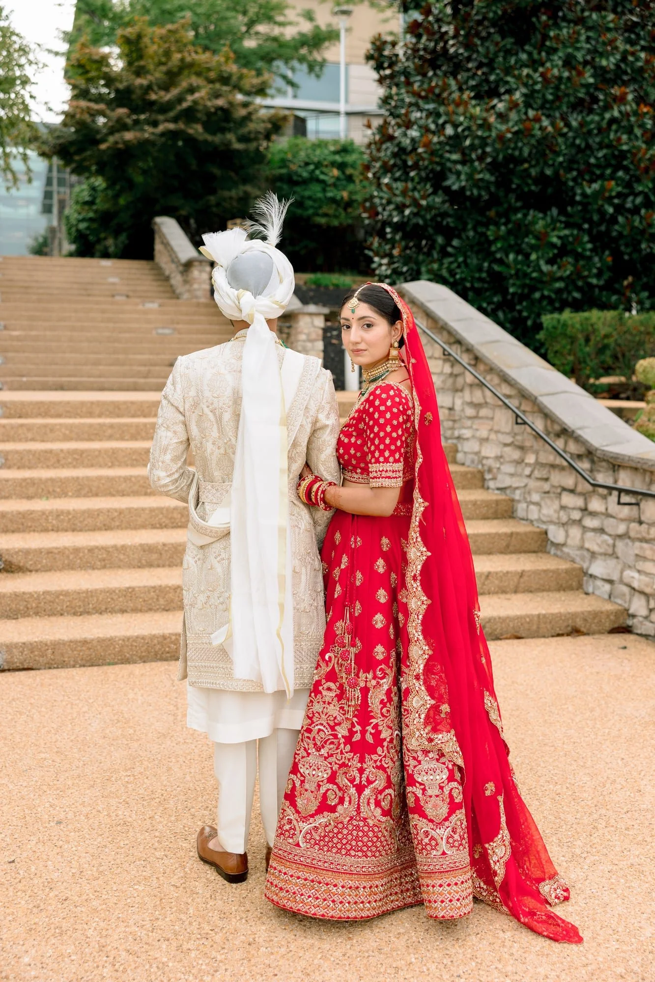 A couple dressed in traditional Indian wedding attire standing outdoors on a staircase among trees. The woman is wearing a red embroidered bridal outfit with gold jewelry, and the man is dressed in a cream-colored sherwani with a turban, seen from be
