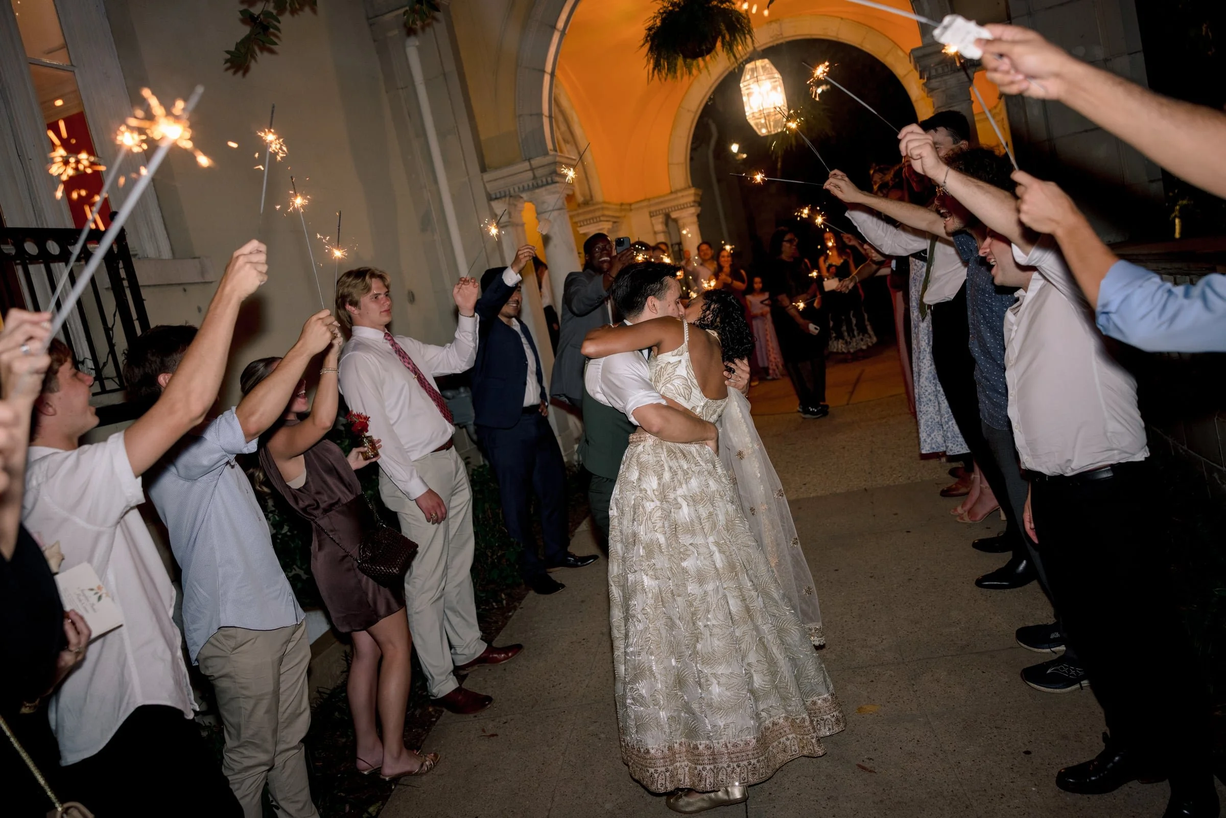 People celebrating at night with sparklers in front of a building with an arch and hanging lights, embracing and smiling.