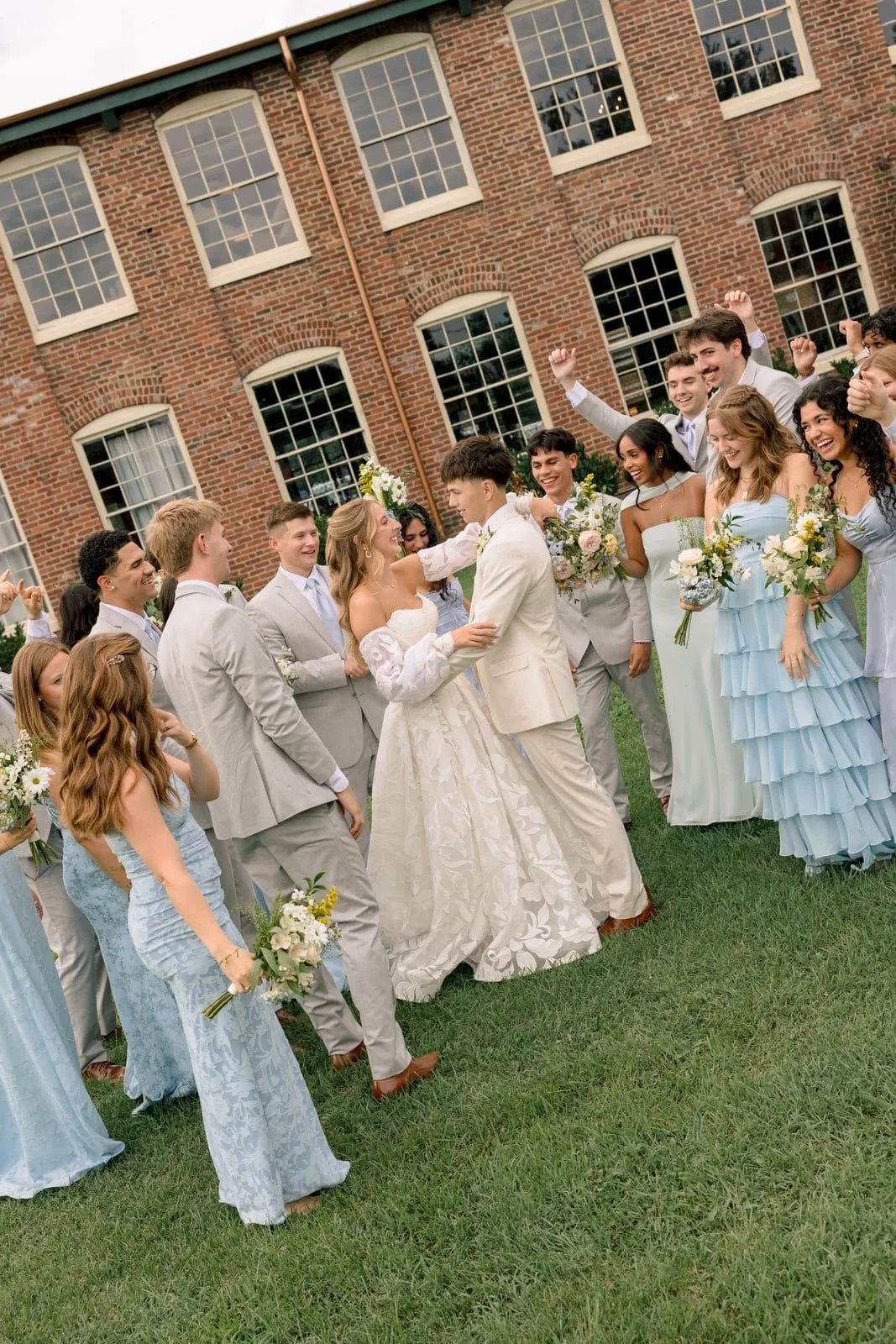 A group of people, including a bride and groom, celebrating outdoors in front of a brick building. The bride and groom are holding hands and smiling, surrounded by friends dressed in formal attire, some holding bouquets of flowers.