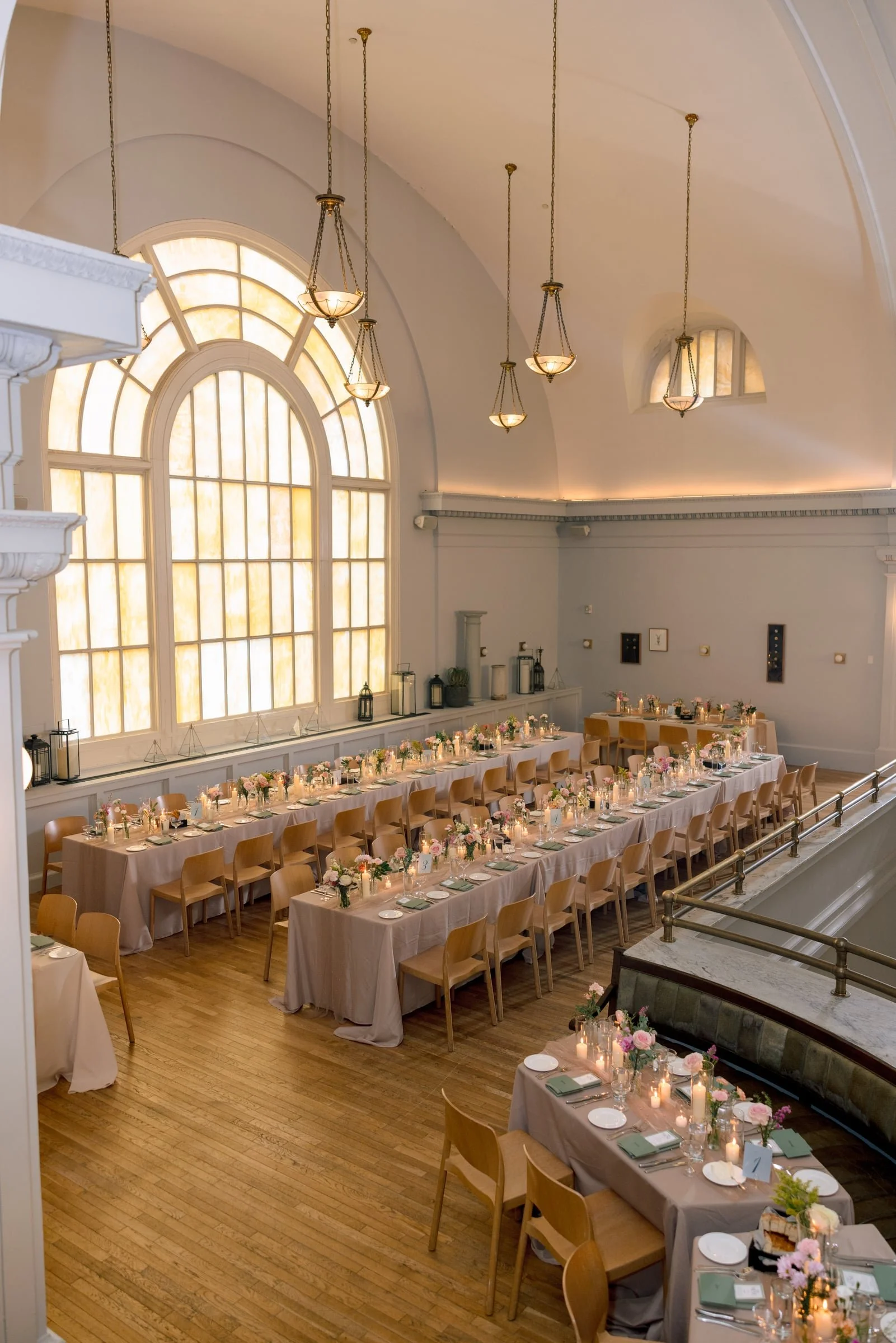 Elegant banquet hall with high arched windows, hanging chandeliers, and long tables decorated with floral centerpieces, candles, and place settings for a formal event or celebration.