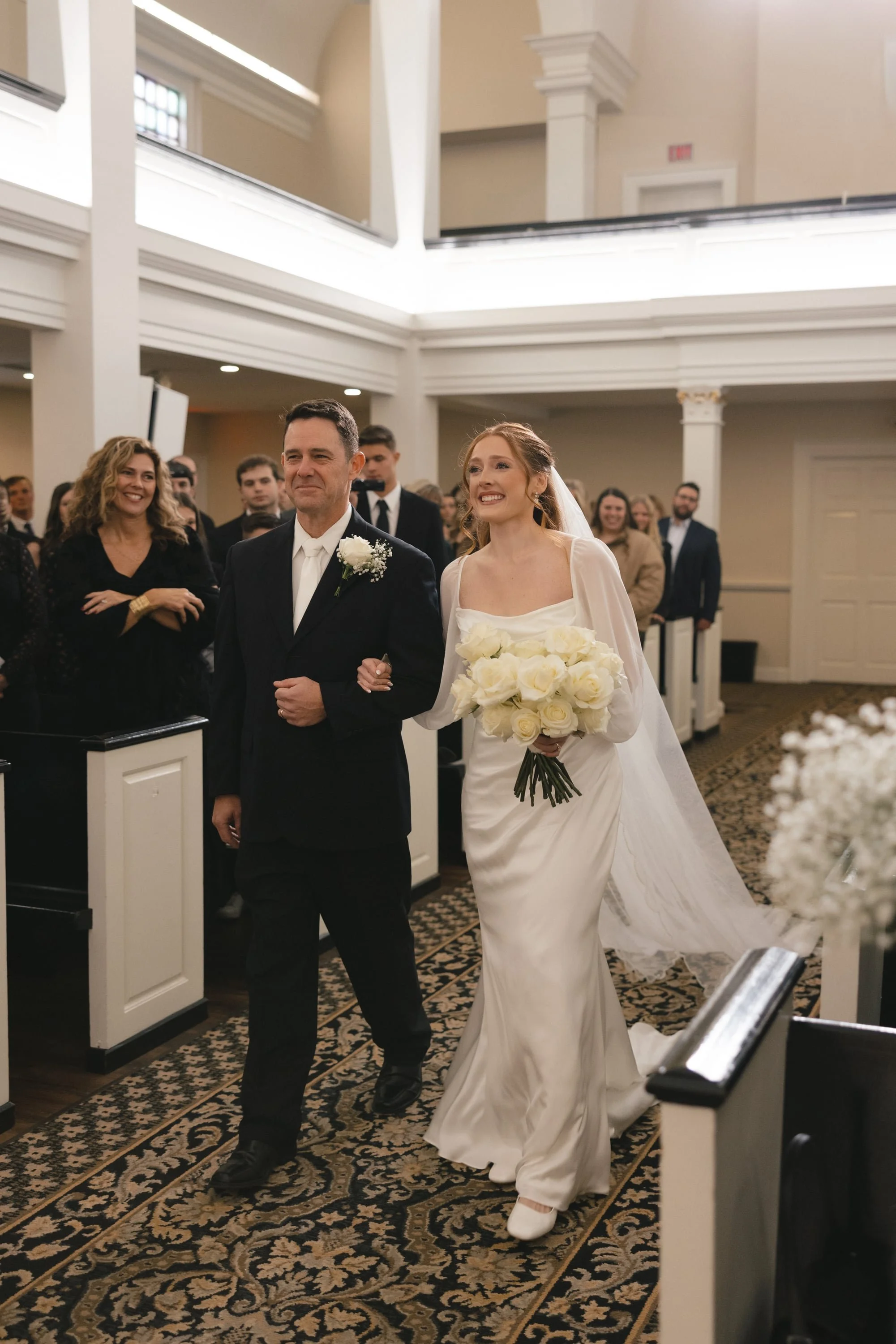 A bride in a white wedding dress holding a bouquet of white roses walks down the aisle, escorted by a man in a black suit and white shirt during a wedding ceremony inside a church, with guests watching and smiling.