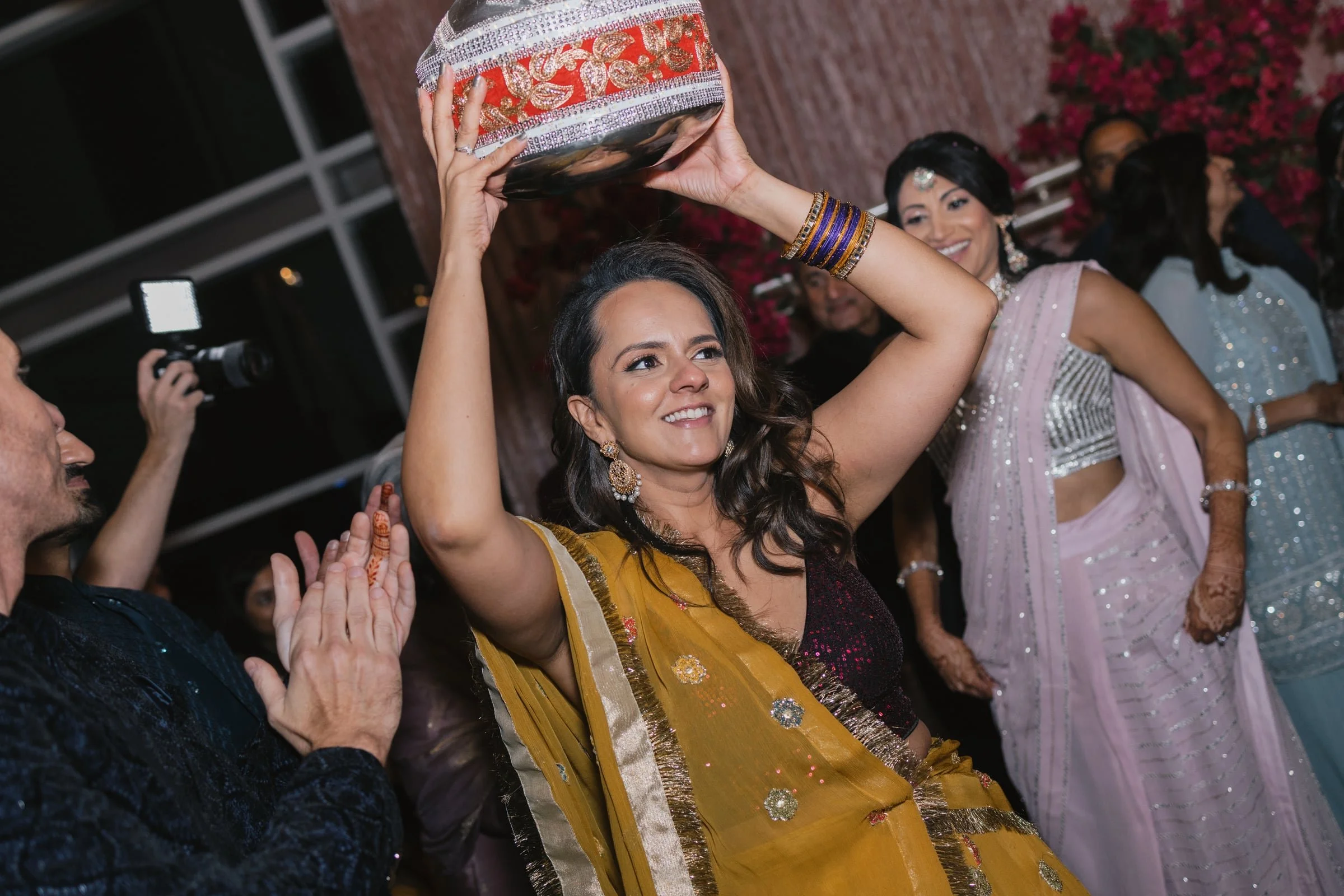 Women dressed in traditional Indian attire enjoying a celebration, with one woman holding a decorated object above her head.