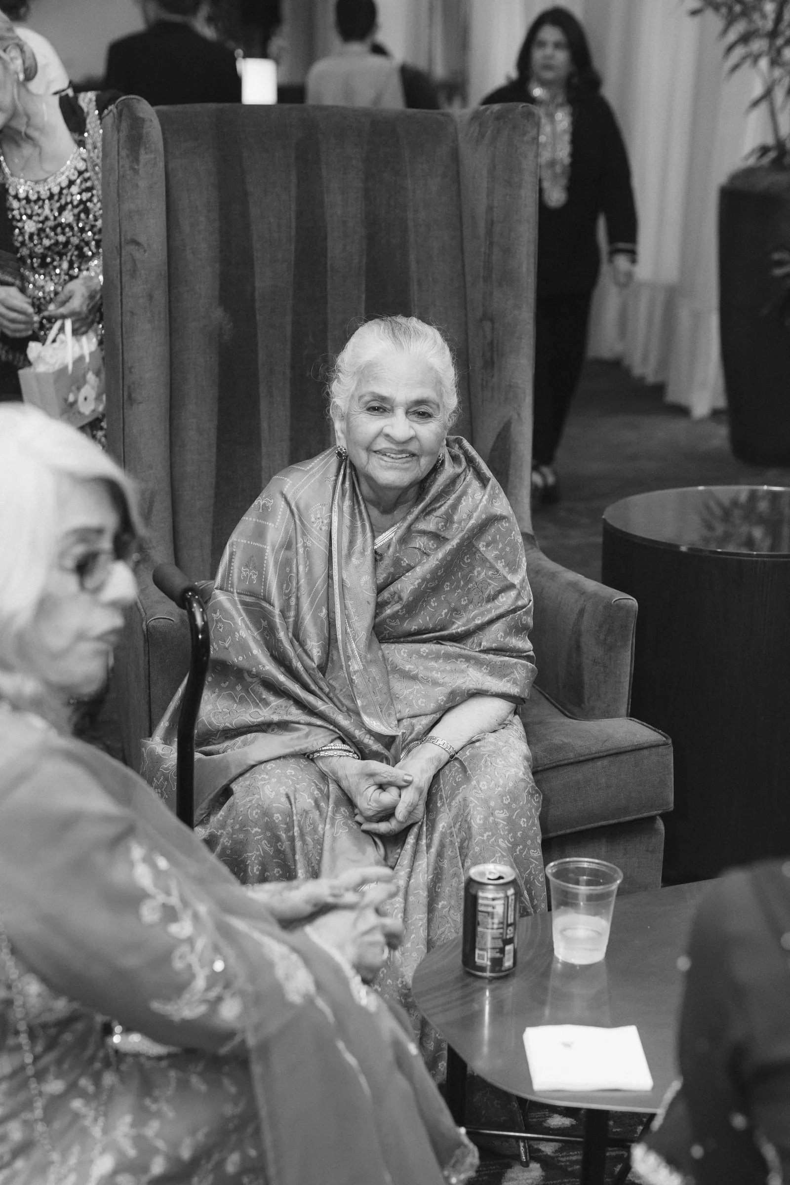 An elderly woman with white hair and a patterned silk outfit sitting on a cushioned chair, smiling, with her hands clasped in her lap. She is at a social gathering, with other people and tables visible in the background.
