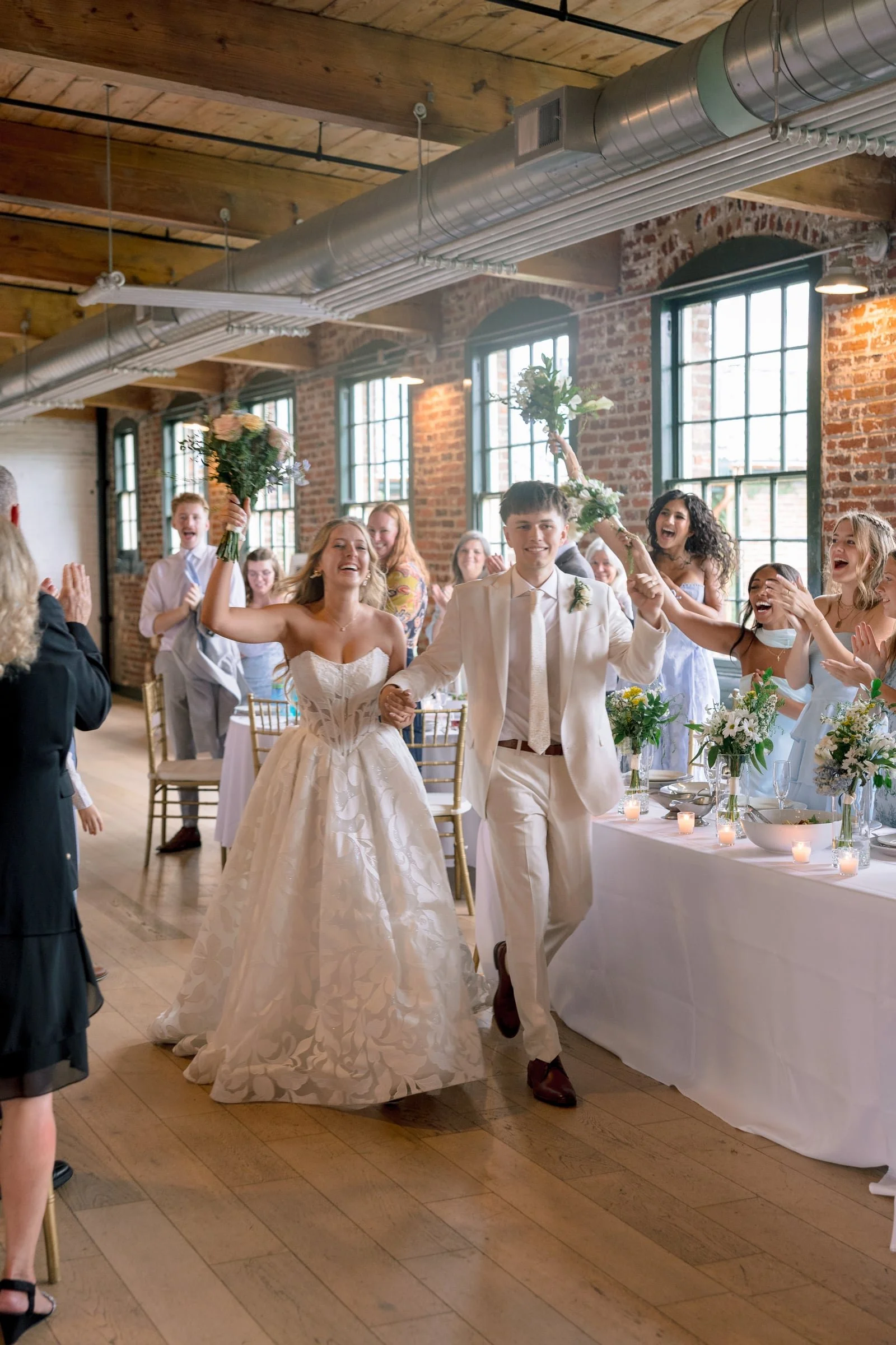 A newlywed couple walking into their wedding reception inside a rustic venue with exposed brick walls and large windows. The bride is holding a bouquet in the air, and the groom is smiling. Guests are celebrating around them, clapping and cheering.