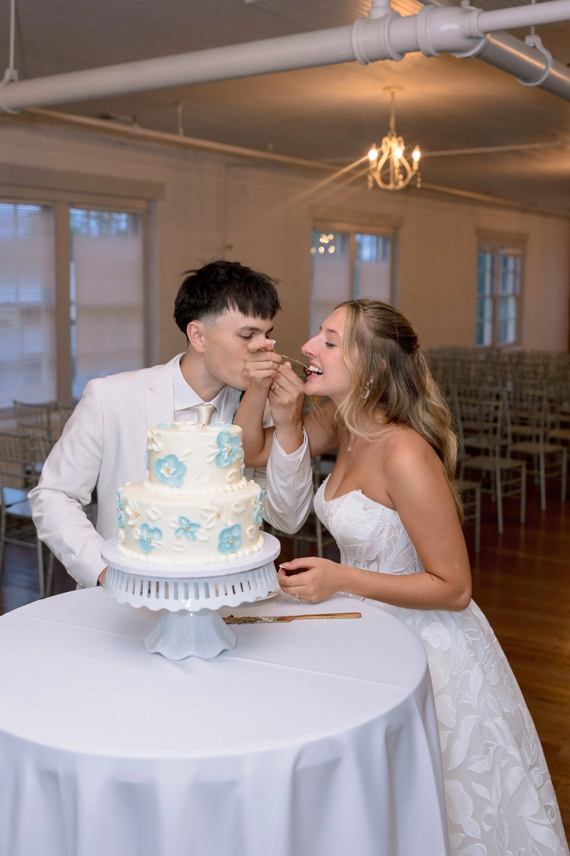 A bride and groom in wedding attire feeding each other cake at their wedding reception. A white wedding cake decorated with blue flowers is on a table in front of them.