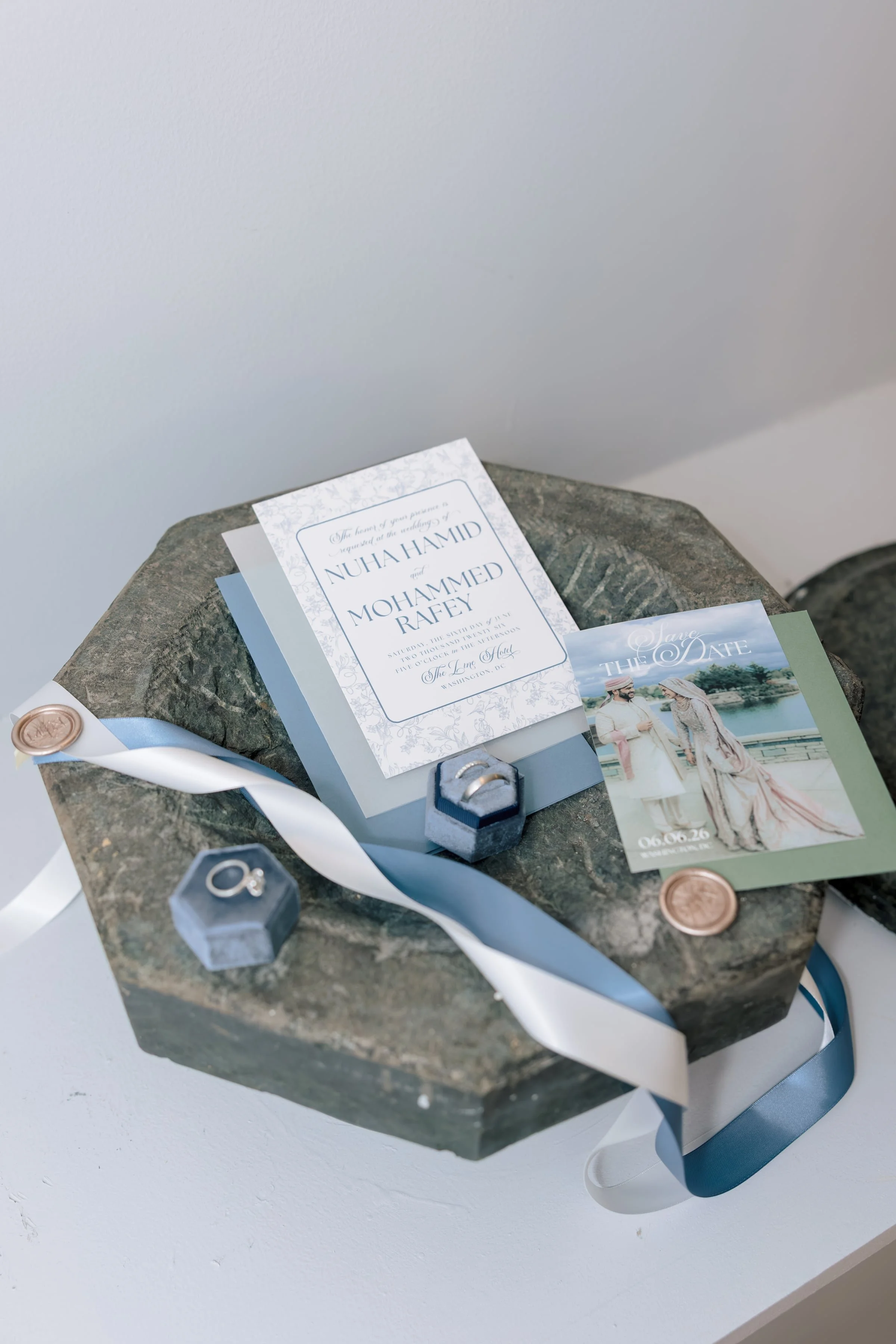 Wedding invitation, save the date card, and wedding rings on a stone surface with wedding-themed decorations.