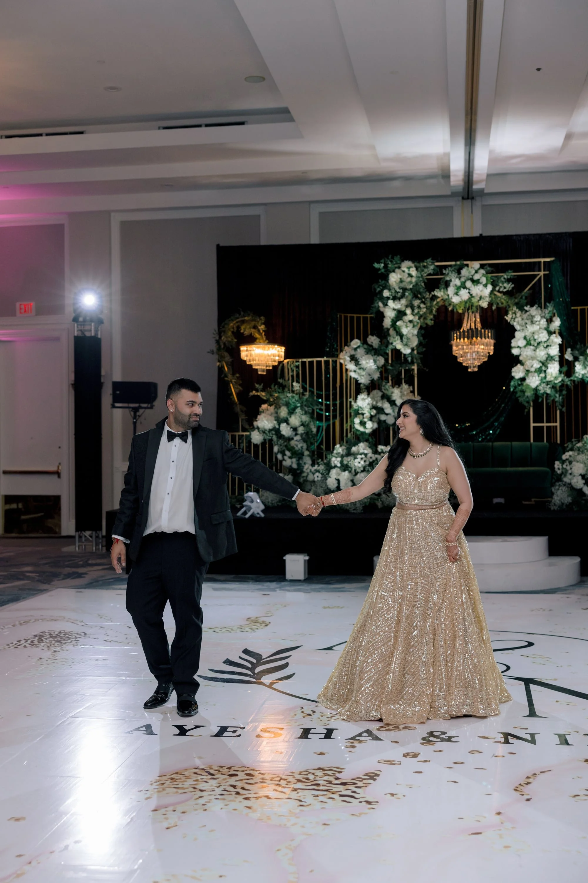 A couple dressed in formal attire dancing at a wedding reception with a decorated stage in the background.