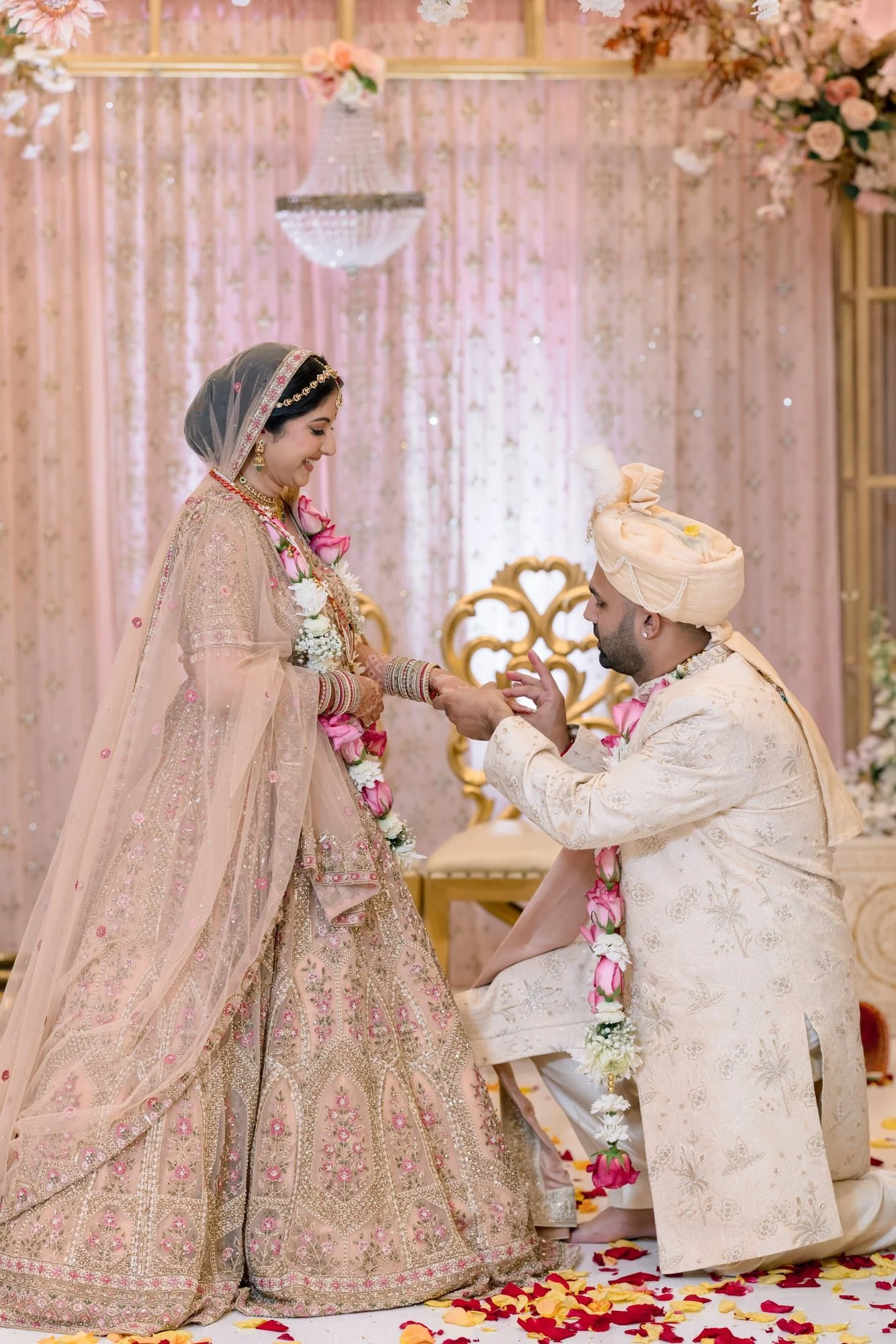 A bride and groom participating in a traditional wedding ceremony. The bride is wearing an ornate pink and gold embroidered dress with jewelry, and the groom is dressed in a cream sherwani with a turban. The bride is placing a ring on the groom's fin