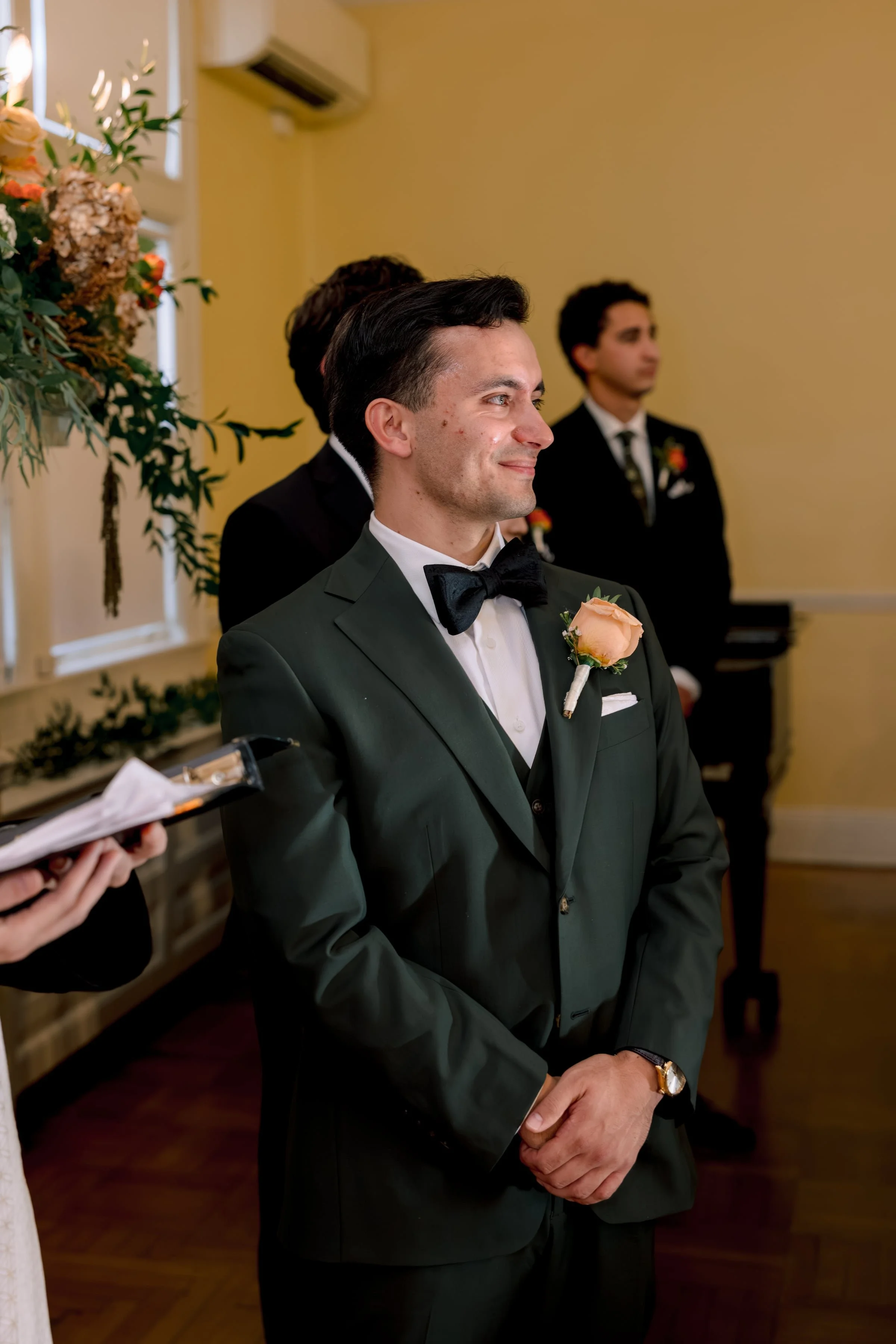 A man dressed in a tuxedo with a black bow tie and a peach-colored boutonniere is attending a formal event, standing with his hands clasped in front of him, smiling softly. Behind him, other men in tuxedos and a large floral arrangement are visible.