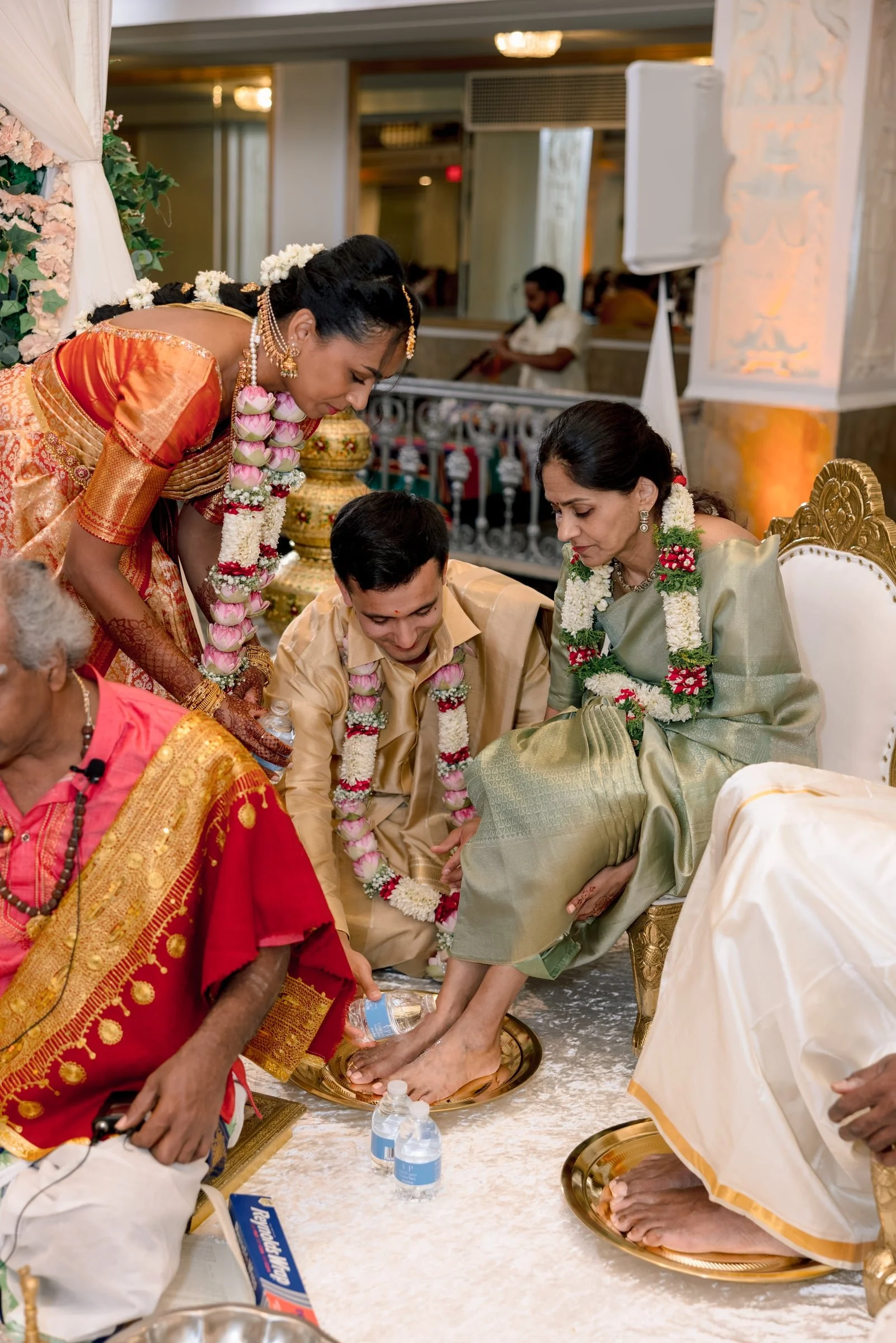 Indian wedding ceremony with women in traditional attire and flower garlands, performing a ritual involving water and feet.