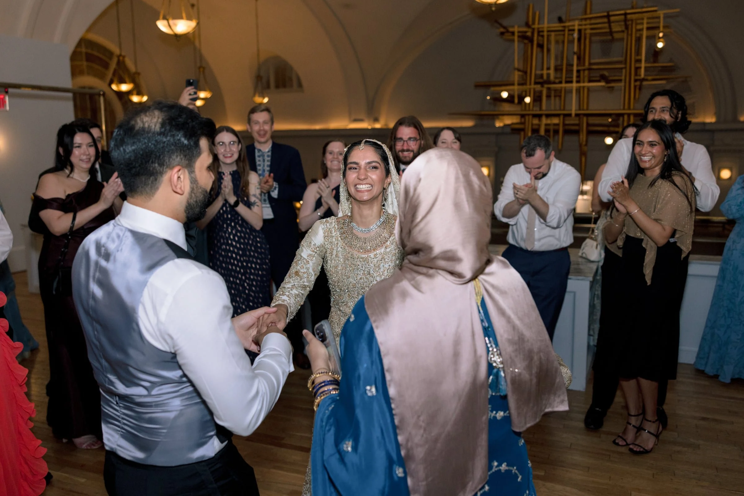 A group of people at a wedding celebration dancing and smiling. The bride and a guest are holding hands, with guests clapping and enjoying the moment indoors with elegant lighting.