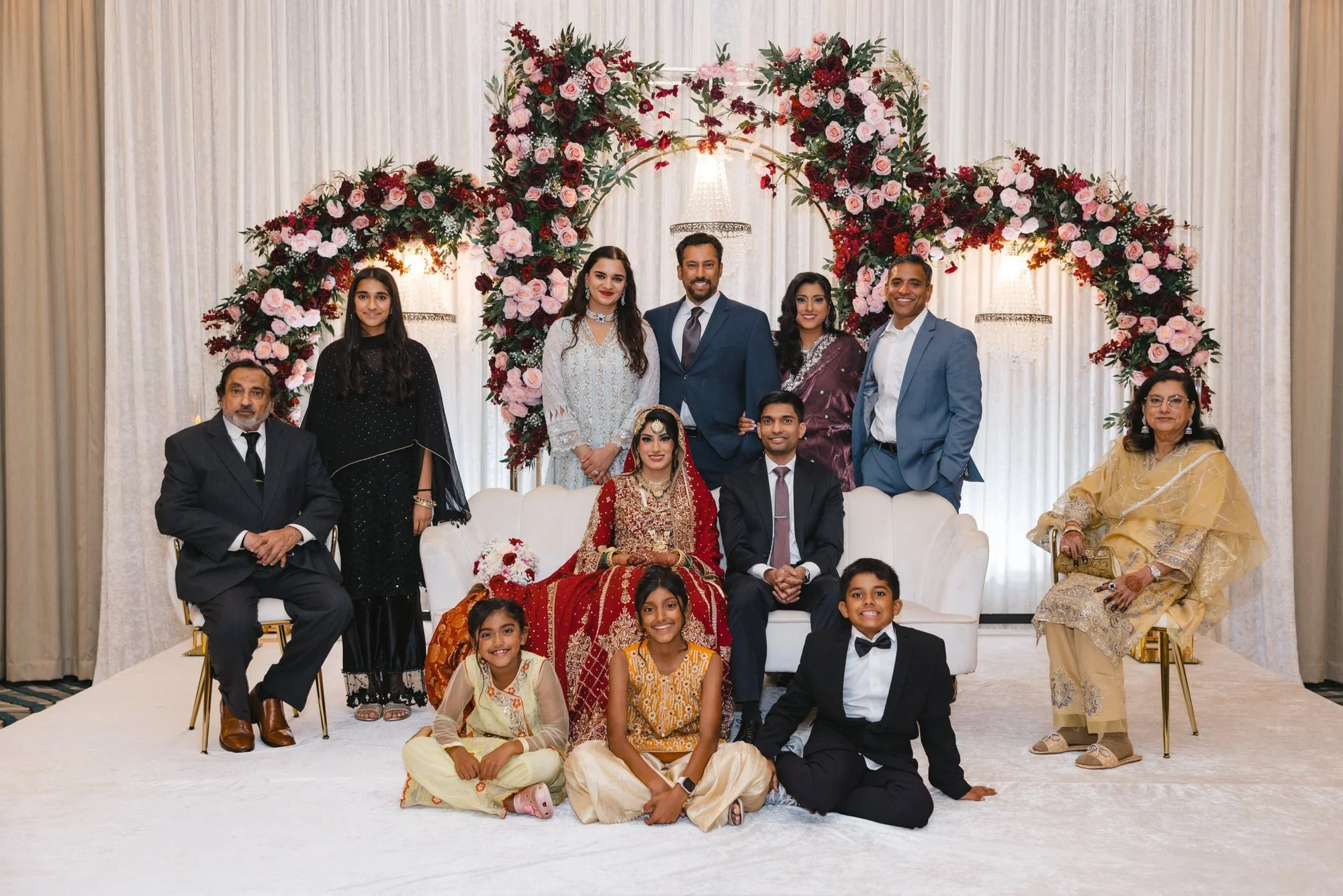 A large family gathers for a group photo at a celebration, with members dressed in formal and traditional attire, seated and standing in front of a floral backdrop with pink and red roses and greenery.