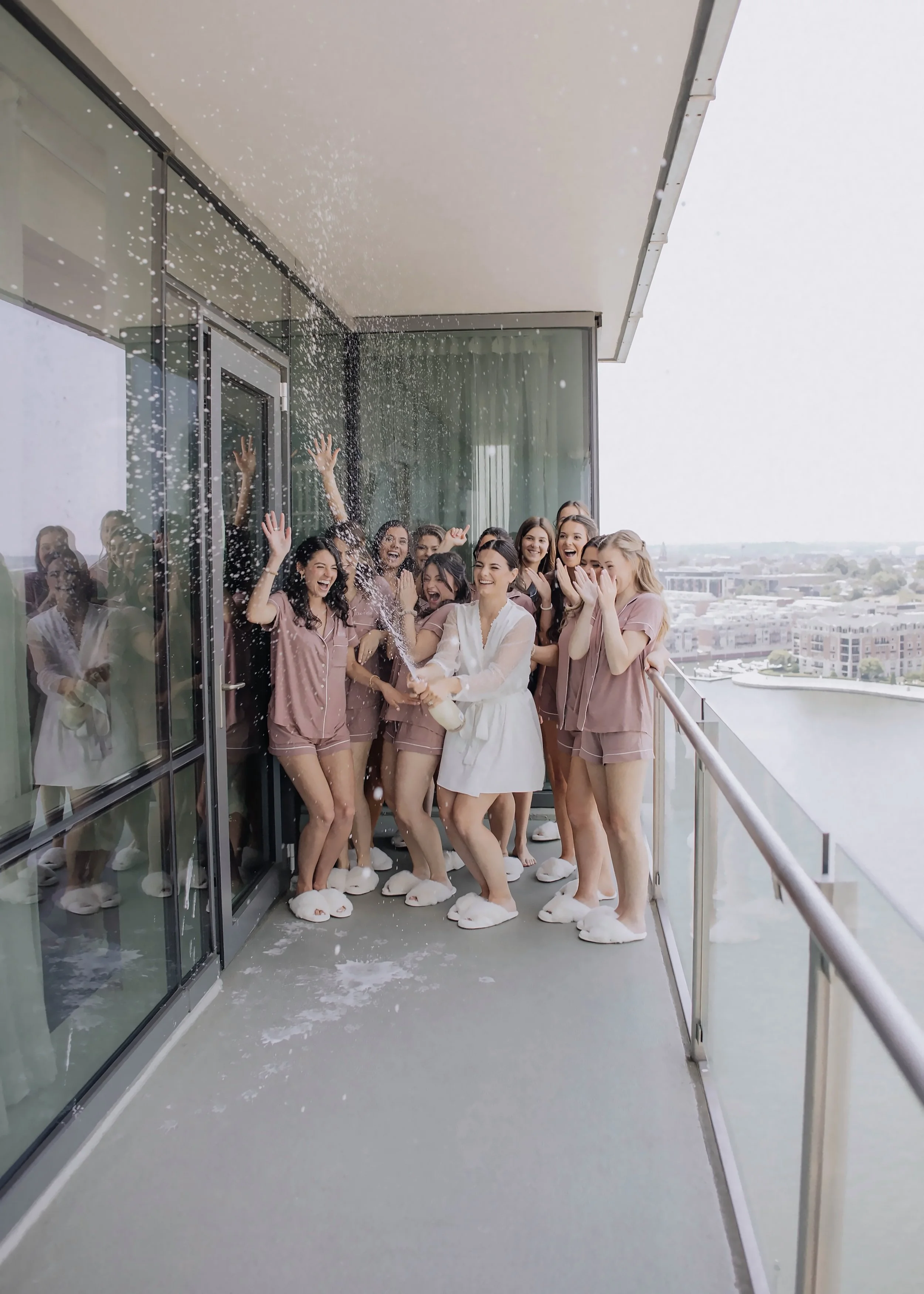 Group of women in pajamas wearing bunny slippers, celebrating on a balcony, with some spraying foam or snow and laughing.