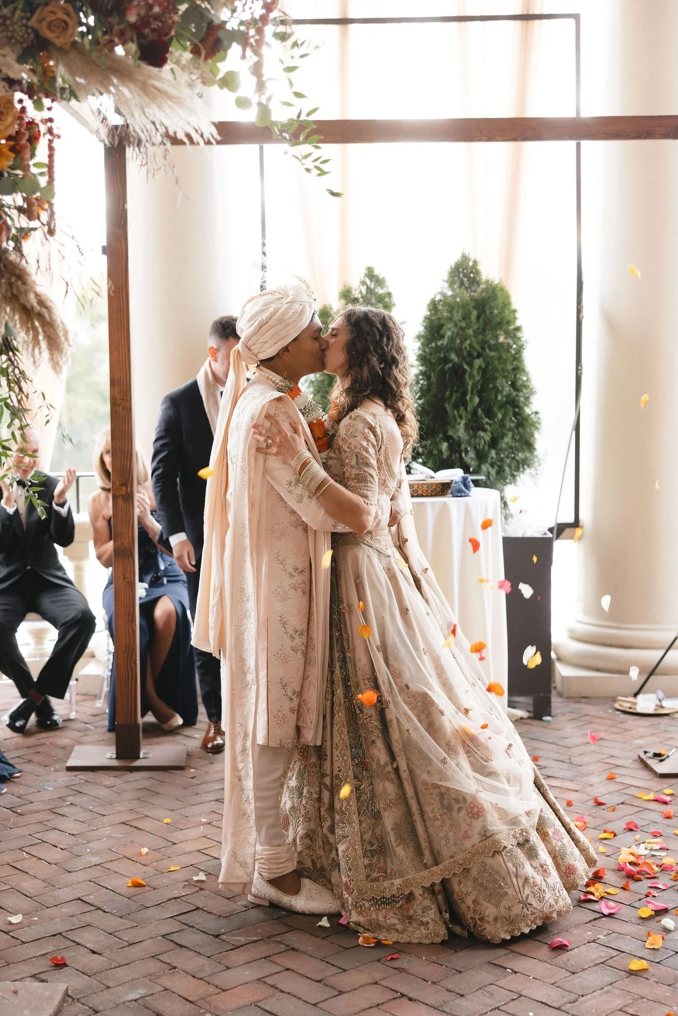 Two women, one wearing a traditional sari and the other in a white bridal gown, share a kiss during a wedding ceremony. There are guests in the background, some seated and others standing, with orange and pink petals scattered on the brick floor.