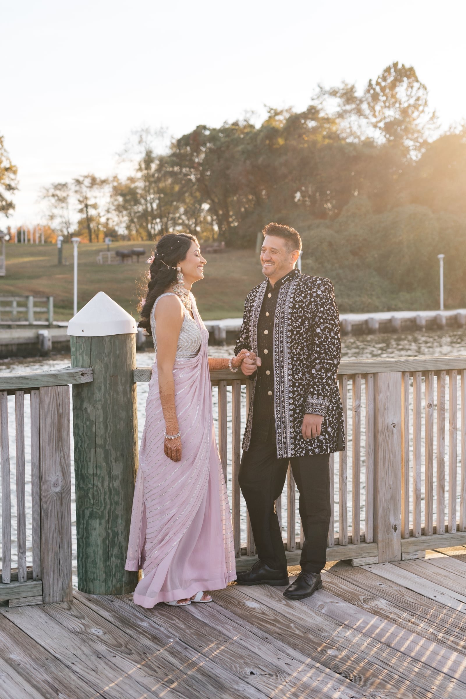 A couple dressed in traditional Indian attire standing on a wooden dock by the water, smiling at each other during sunset.