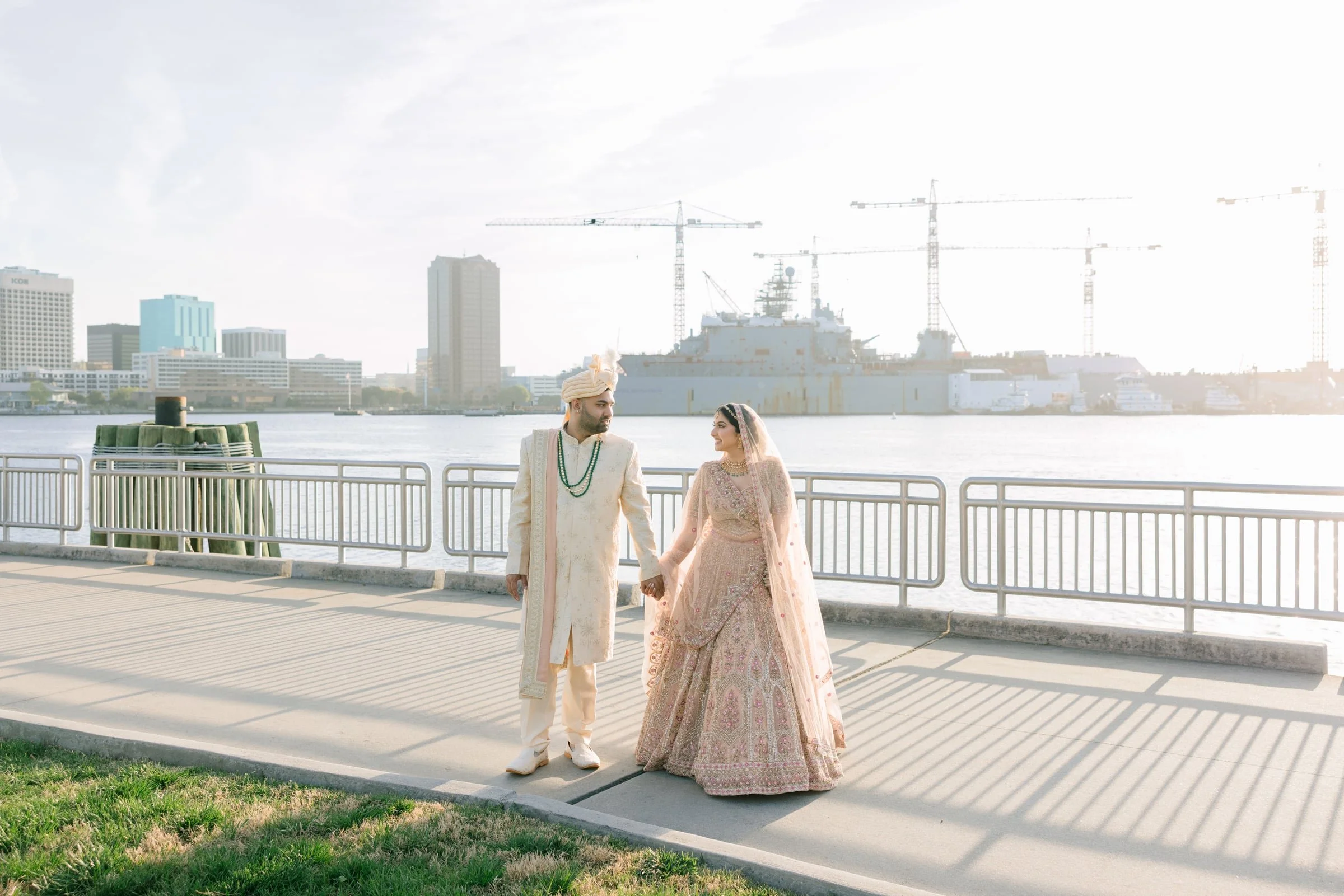 A bride and groom in traditional Indian wedding attire holding hands and walking along a waterfront promenade with city buildings and cranes in the background, during daytime.