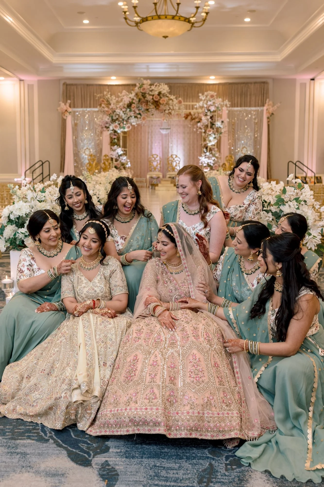A group of women dressed in colorful traditional Indian attire, gathered together in a wedding setting decorated with flowers, smiling and enjoying a celebratory moment.