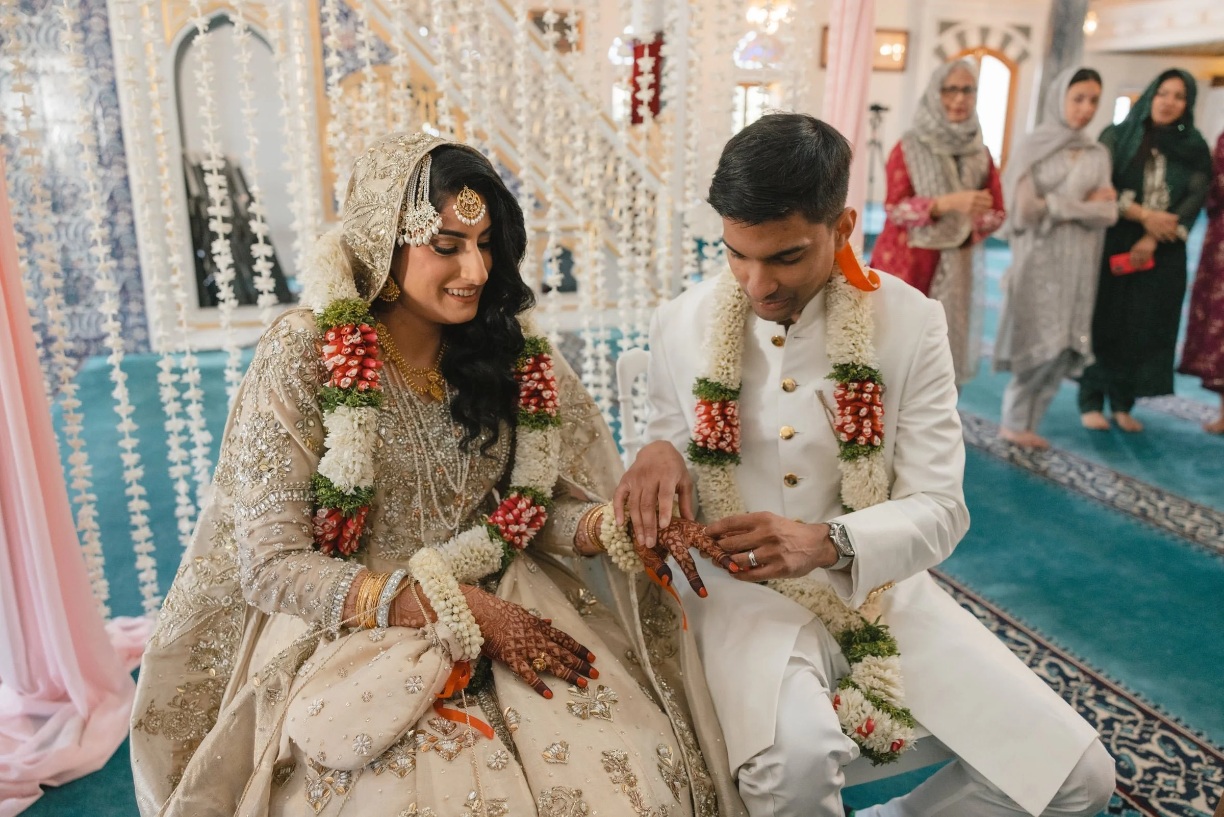 A couple performing a traditional wedding ritual during their wedding ceremony, with women in the background watching.