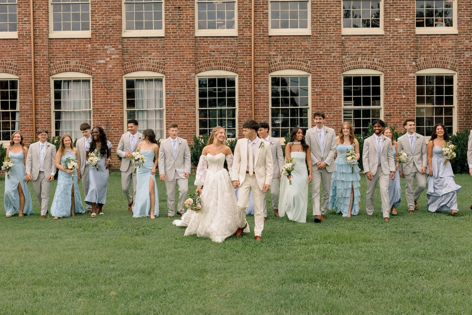 A wedding party posing on a lawn in front of a brick building, with the bride and groom holding hands in the center, surrounded by bridesmaids and groomsmen dressed in formal attire.